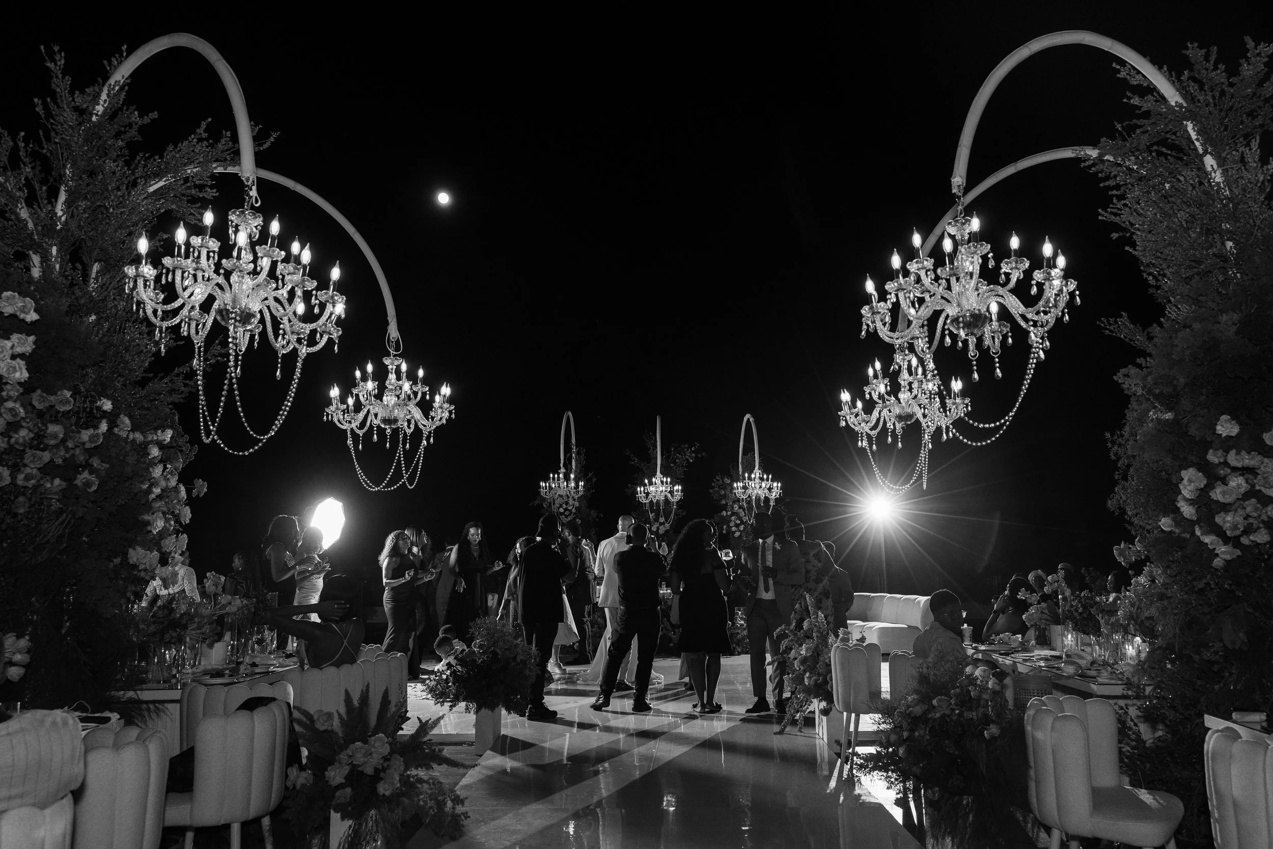 People mingling at an elegant outdoor night event under chandeliers with floral decorations and a full moon in the sky.