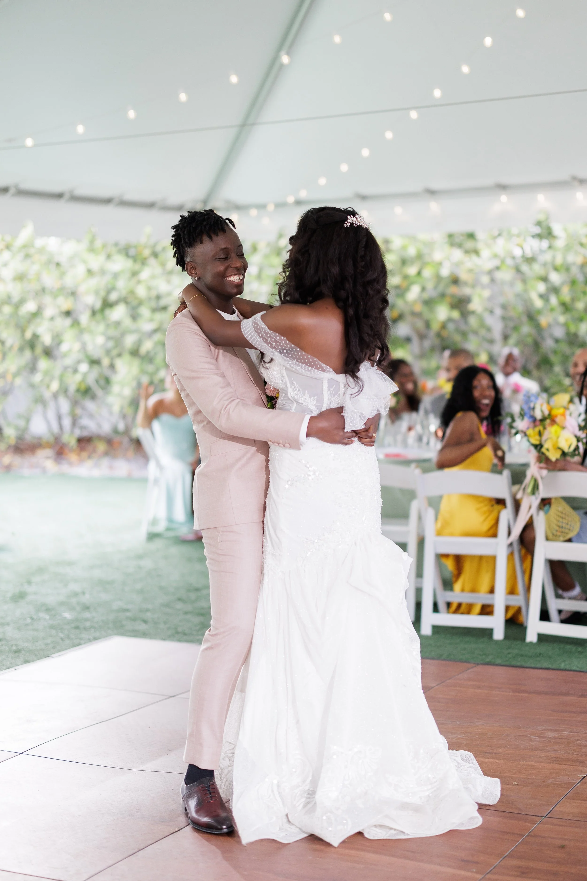 Two women are dancing and hugging at a wedding reception under a white tent, with guests sitting at decorated tables in the background.