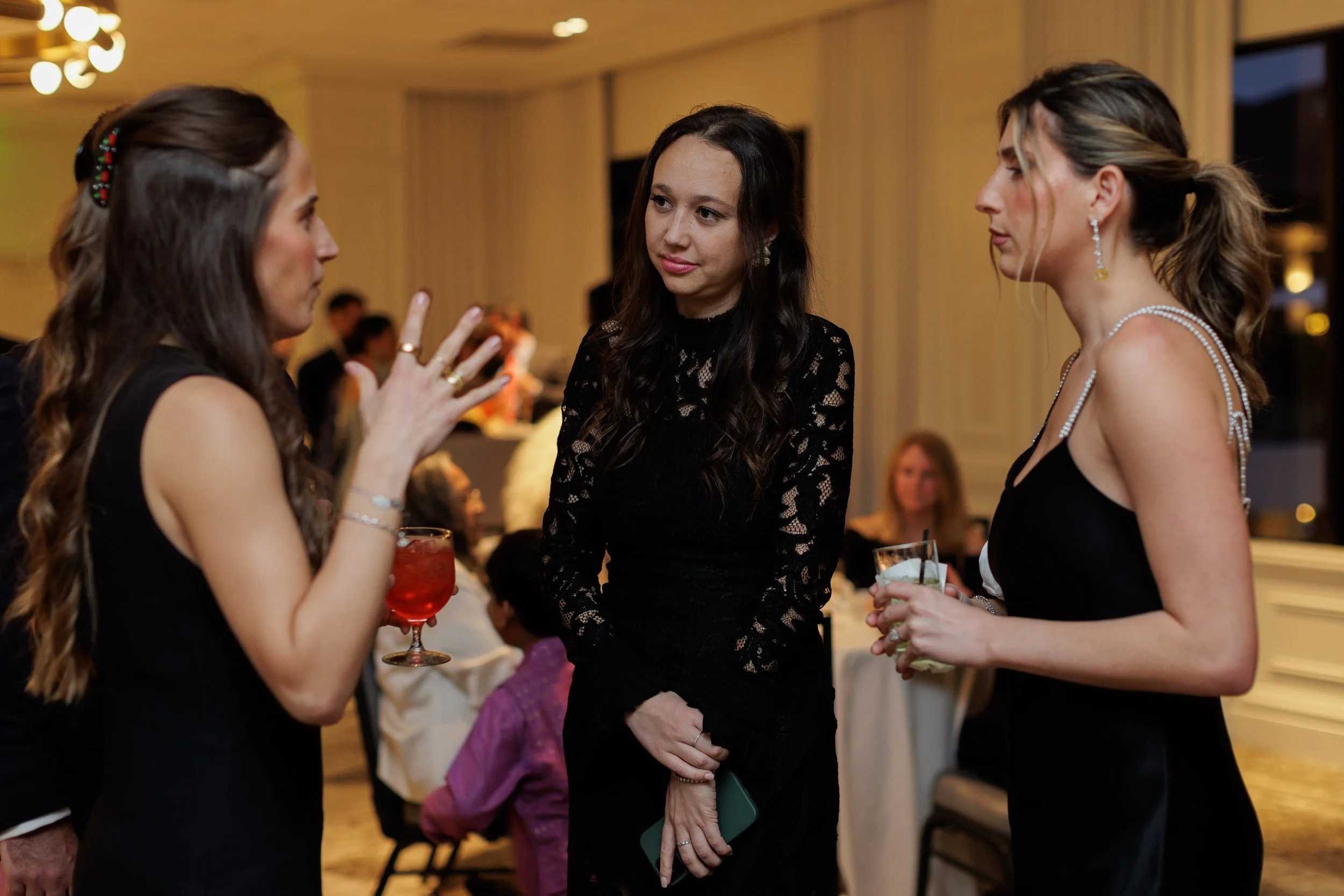 Three women in black dresses engaged in conversation at a social gathering, one holding a red cocktail, another with a glass of clear beverage, and the third holding a smartphone, indoors with dining guests in the background.