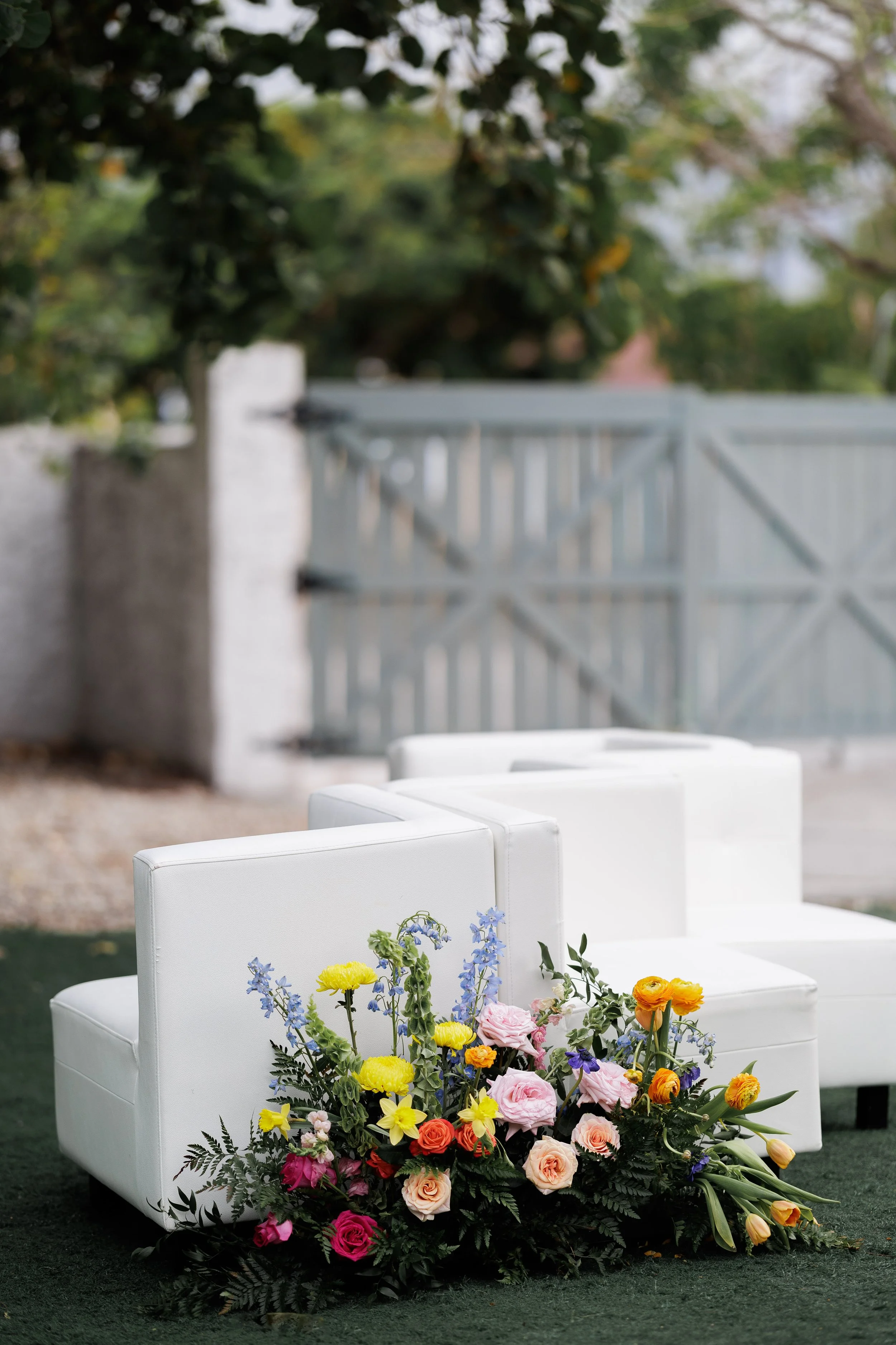 White outdoor chairs arranged outdoors with a colorful floral arrangement at their base, set against a blurred background of a gate and trees.