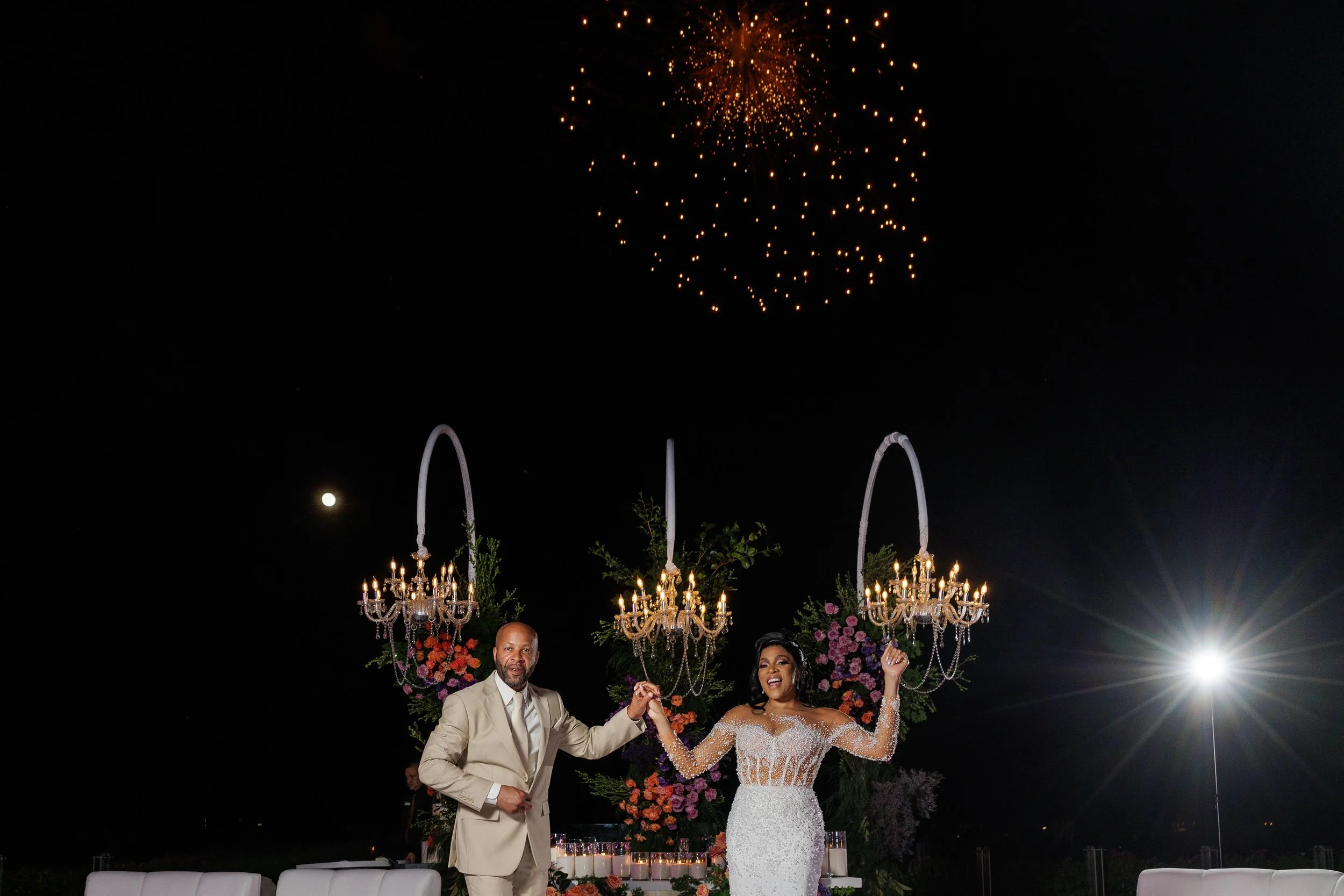 A bride and groom celebrating at their wedding reception outdoors at night, with fireworks in the sky, chandeliers decorating the backdrop, and floral arrangements in the background.