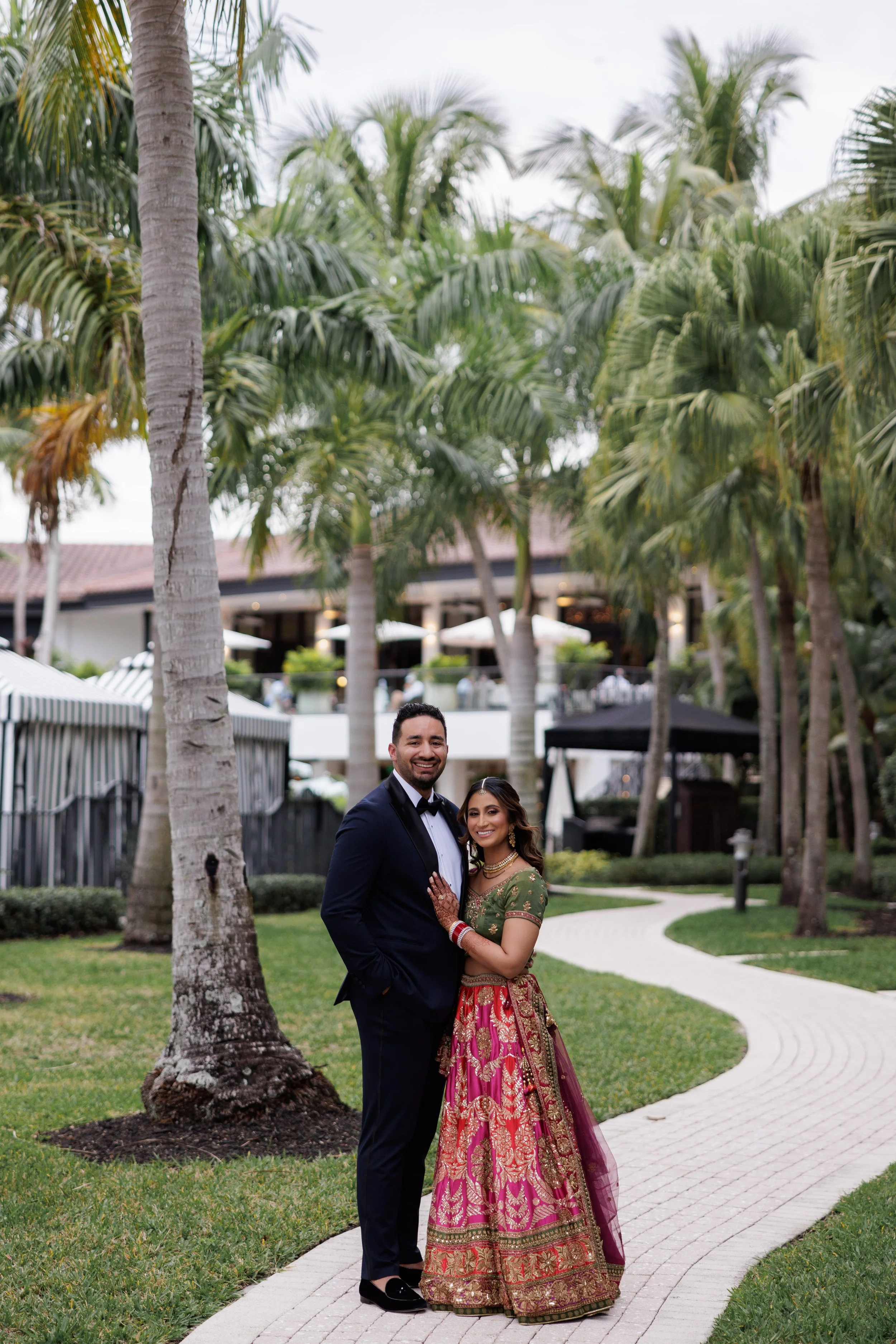 A couple in formal attire, a man in a tuxedo and a woman in traditional Indian dress, standing on a curved pathway among palm trees and lush greenery outdoors.