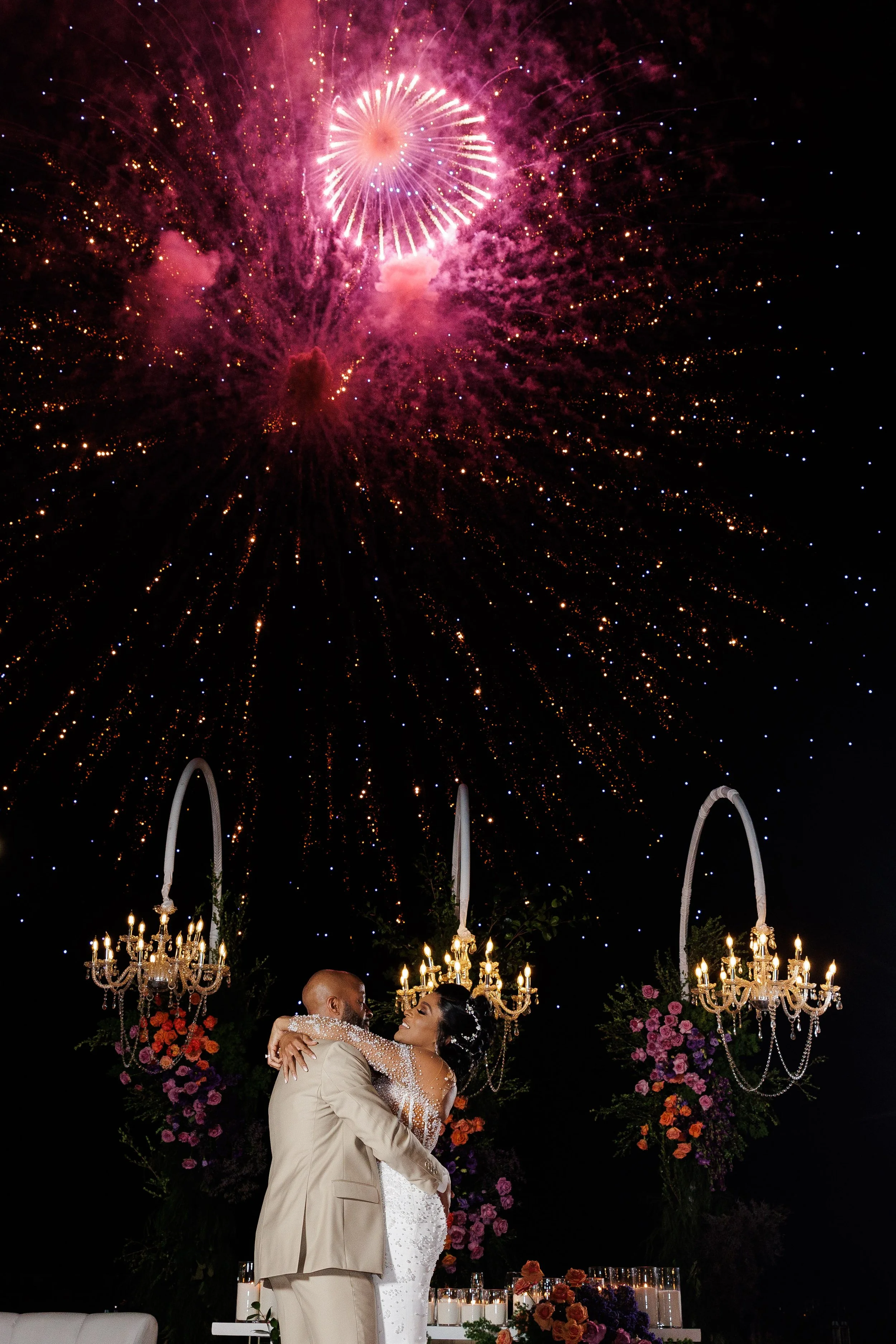 Couple dancing at a wedding reception under fireworks and chandeliers with floral arrangements.