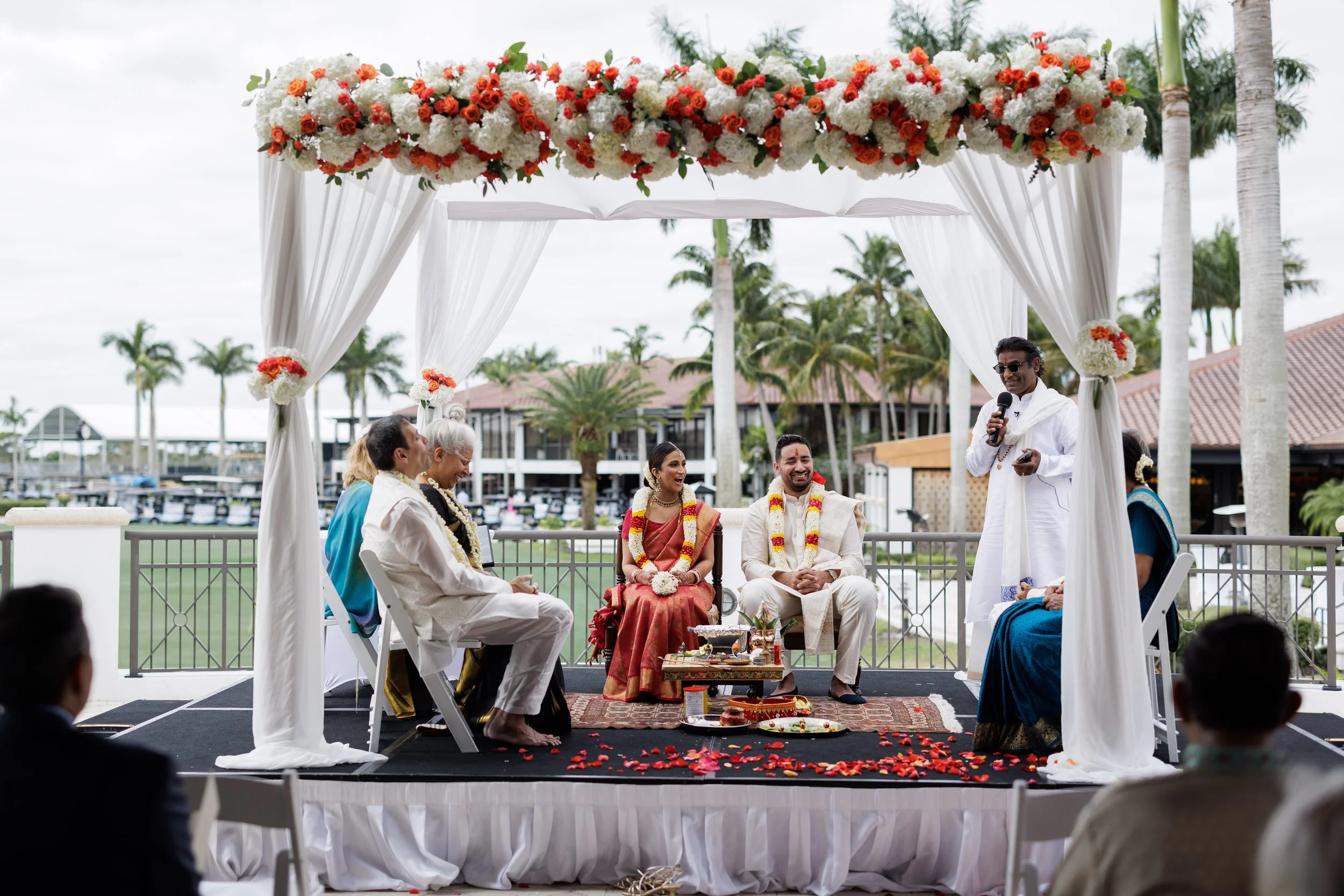 An outdoor wedding ceremony with a decorated floral arch, two brides and two grooms seated on stage, a man in white speaking into a microphone, and guests in the foreground. The setting includes palm trees and water in the background.