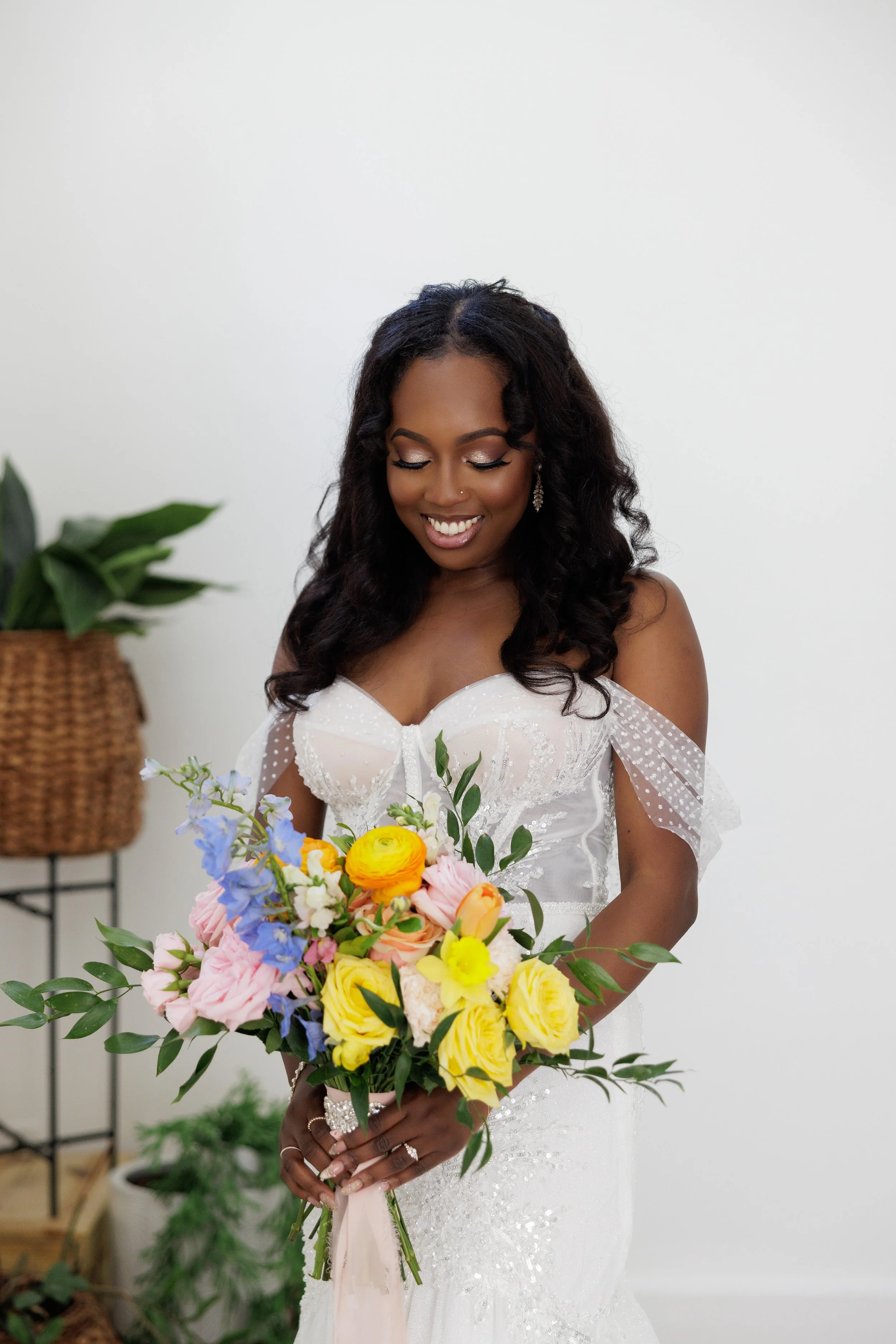 A woman in a white wedding dress holding a colorful bouquet of flowers, smiling and looking down.