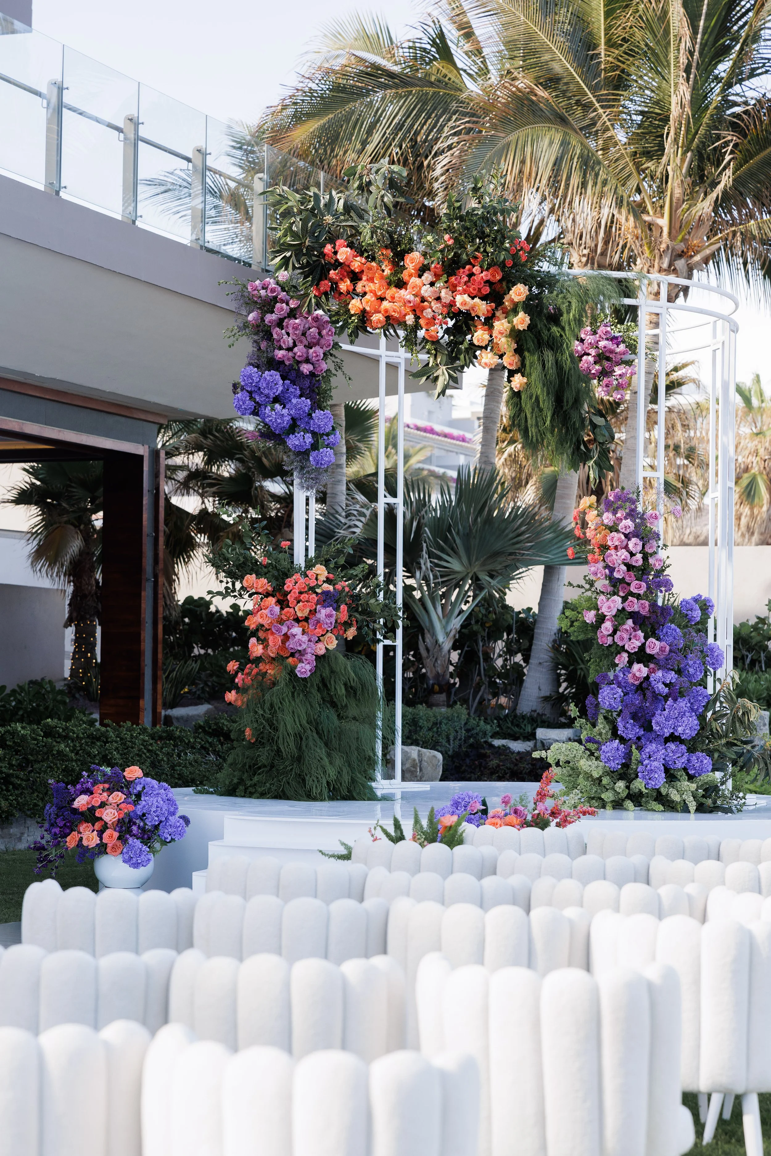 Outdoor wedding ceremony setup with a white arch decorated with pink, orange, purple, and blue flowers, surrounded by palm trees, white chairs, and lush greenery.
