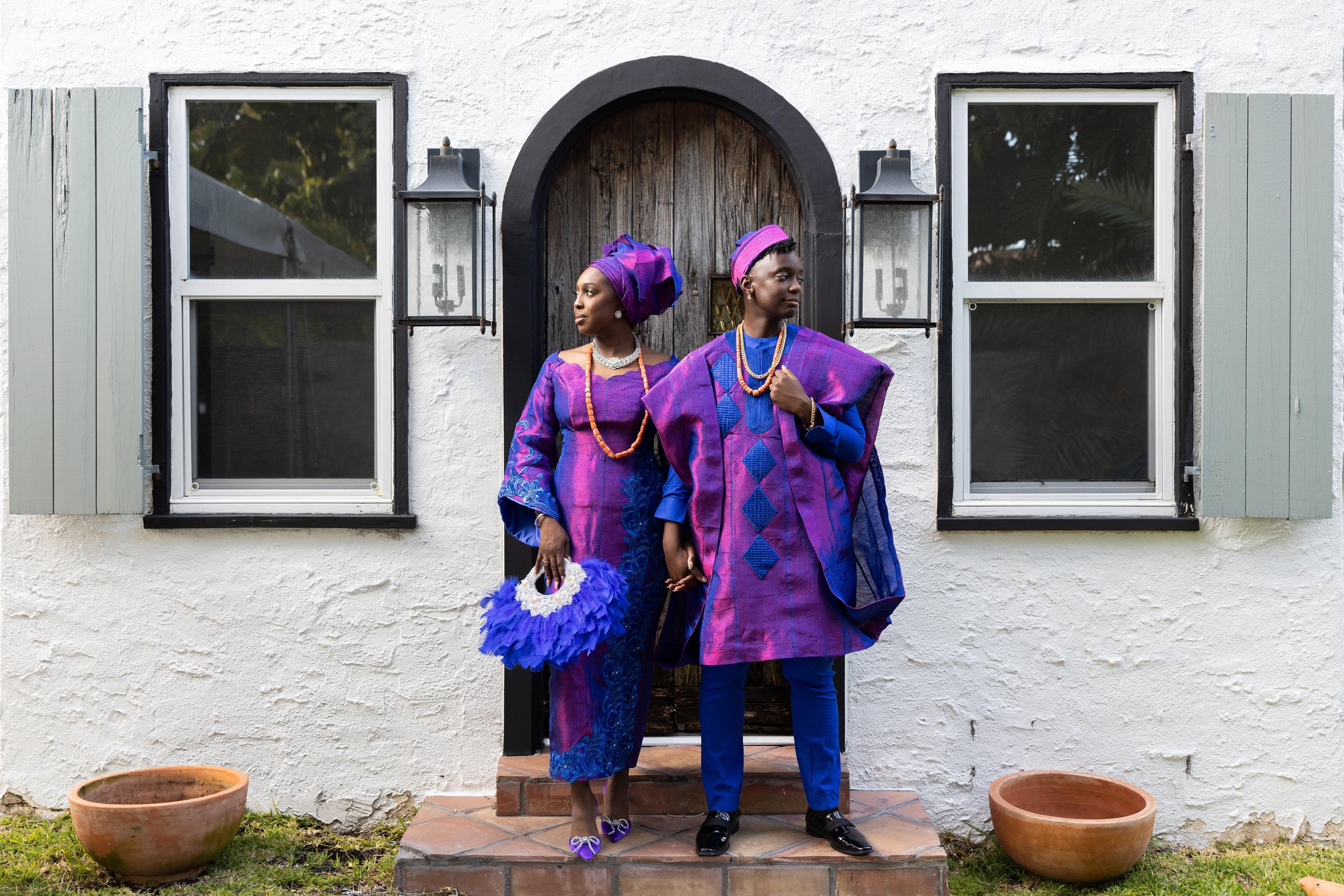 A man and woman of African descent dressed in traditional purple and blue African attire, standing on a brick step in front of a white house with black trim and two windows with wooden shutters, holding hands and posing for the photo.