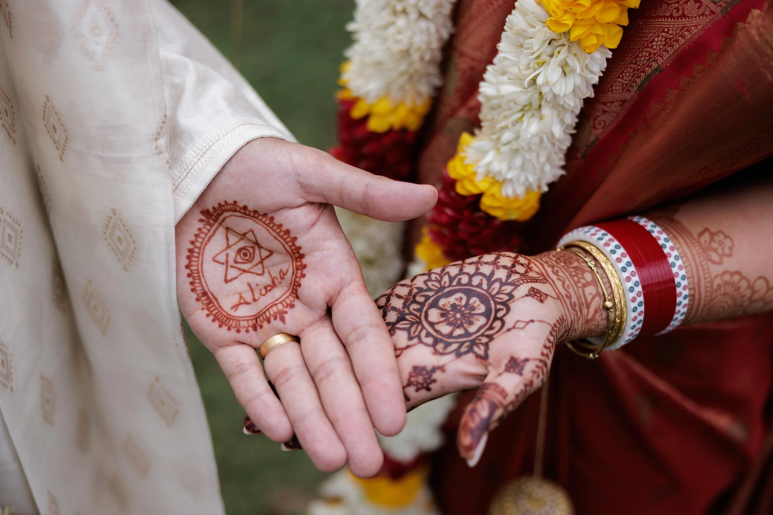 Close-up of a married couple holding hands, showcasing henna tattoos on their hands and wrists, with wedding attire and floral garlands.