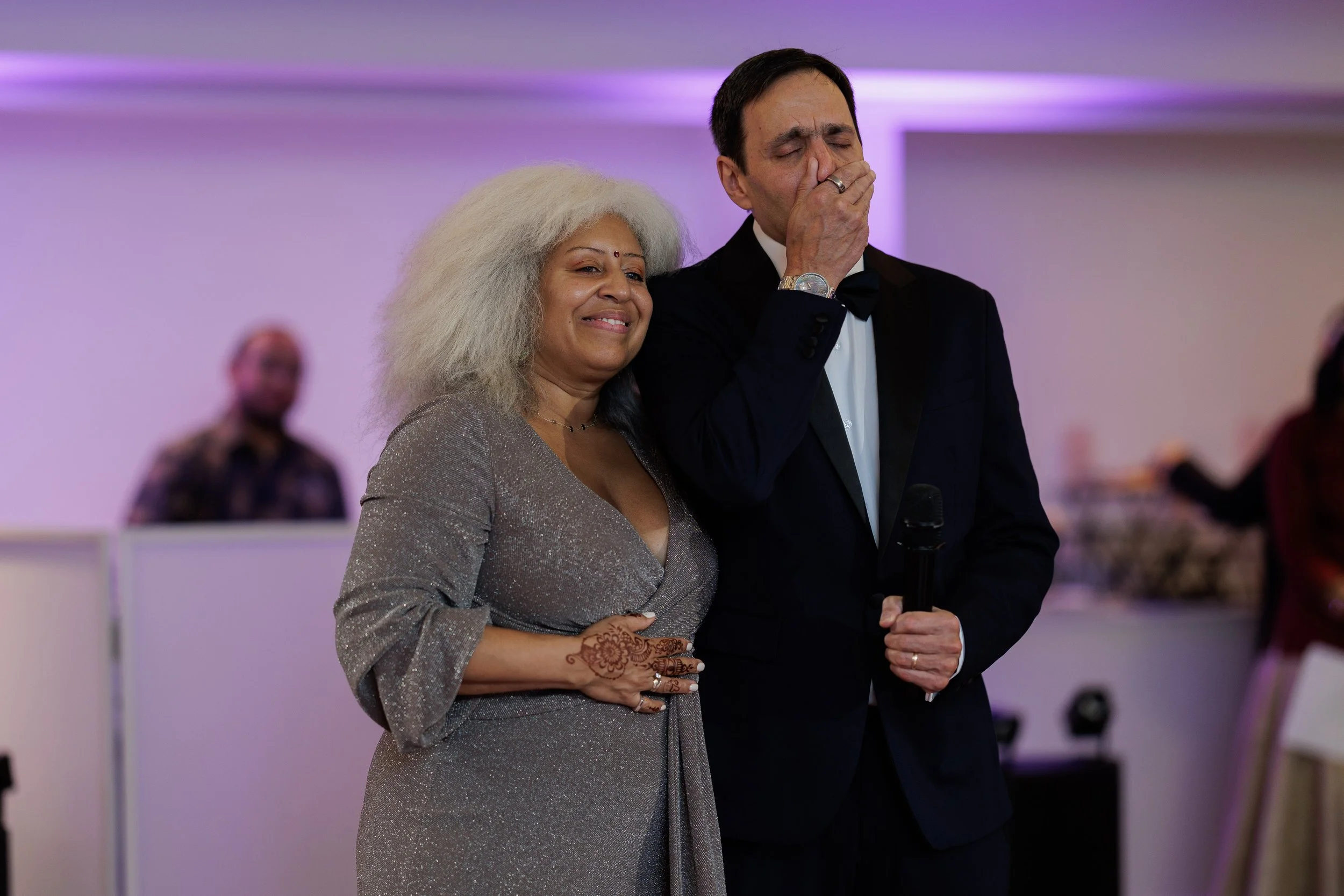 A woman with gray hair and a glittery dress stands next to a man in a tuxedo holding a microphone. The woman is smiling, and the man appears emotional, with his hand covering his mouth. The background shows a blurred person and soft purple lighting.