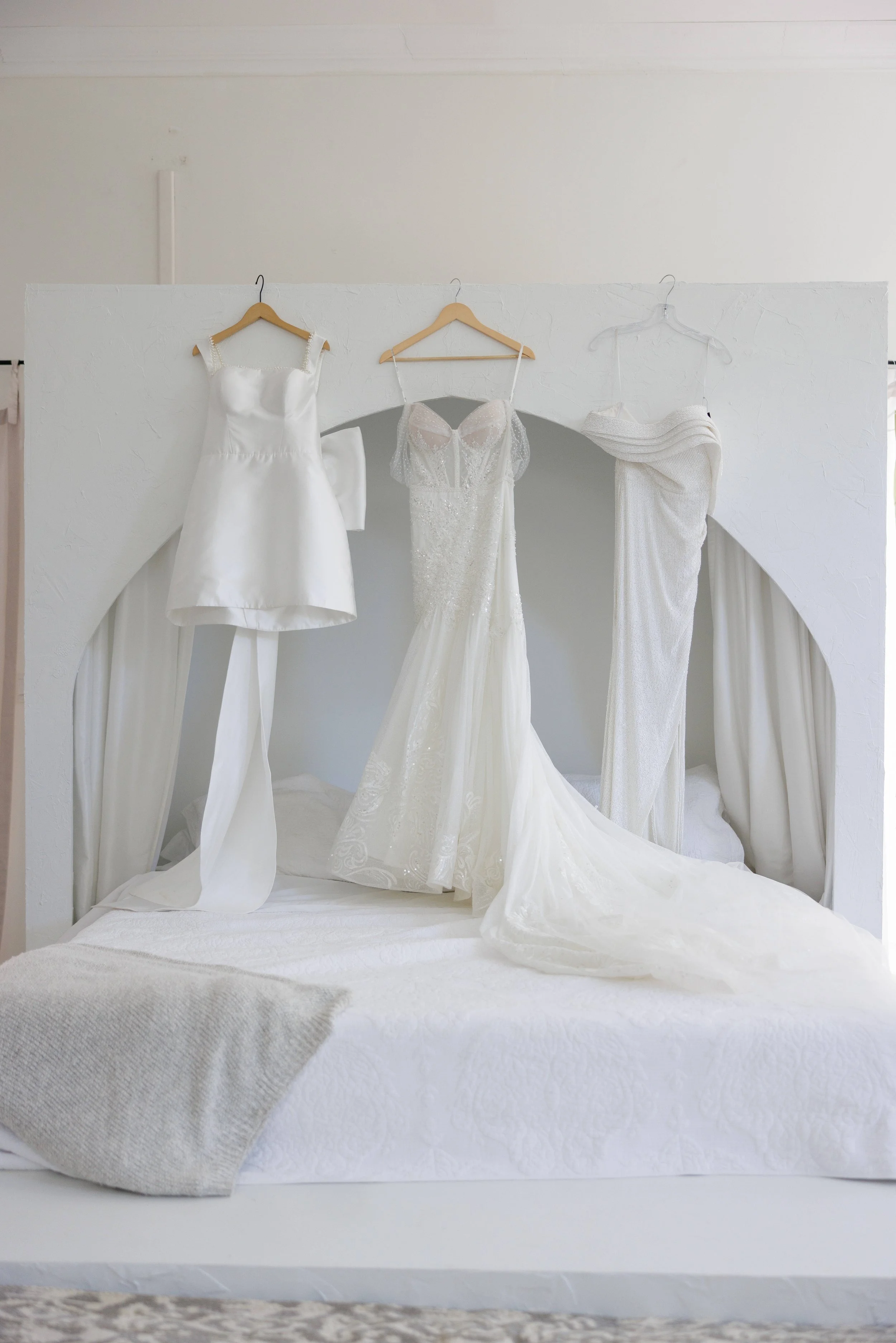 Three wedding dresses hanging on a white bed canopy in a bright room.