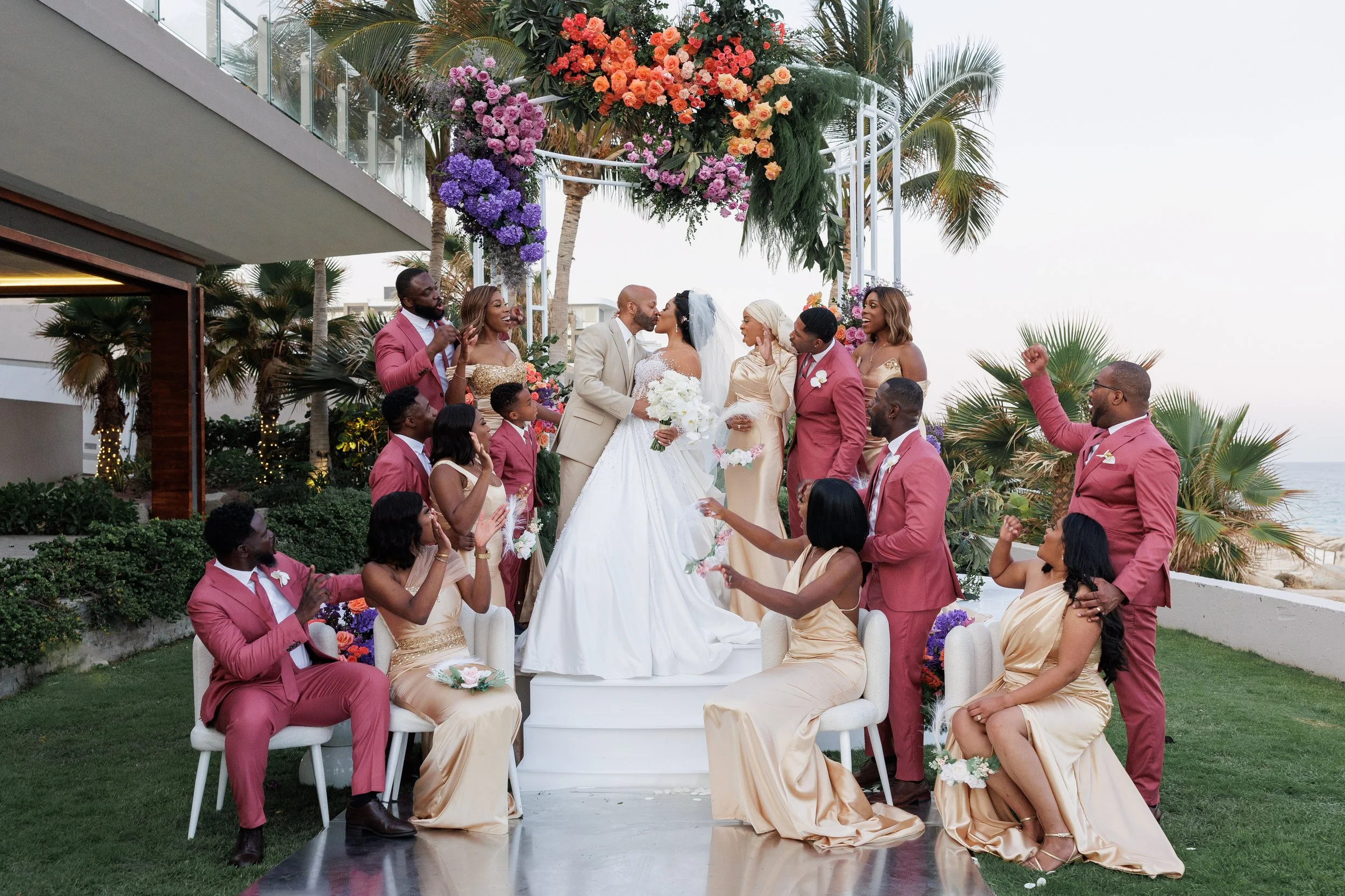 A wedding ceremony on a terrace with a view of the ocean, featuring a bride and groom kissing, surrounded by wedding party members dressed in pink suits and gold dresses, under a floral arch with palm trees in the background.