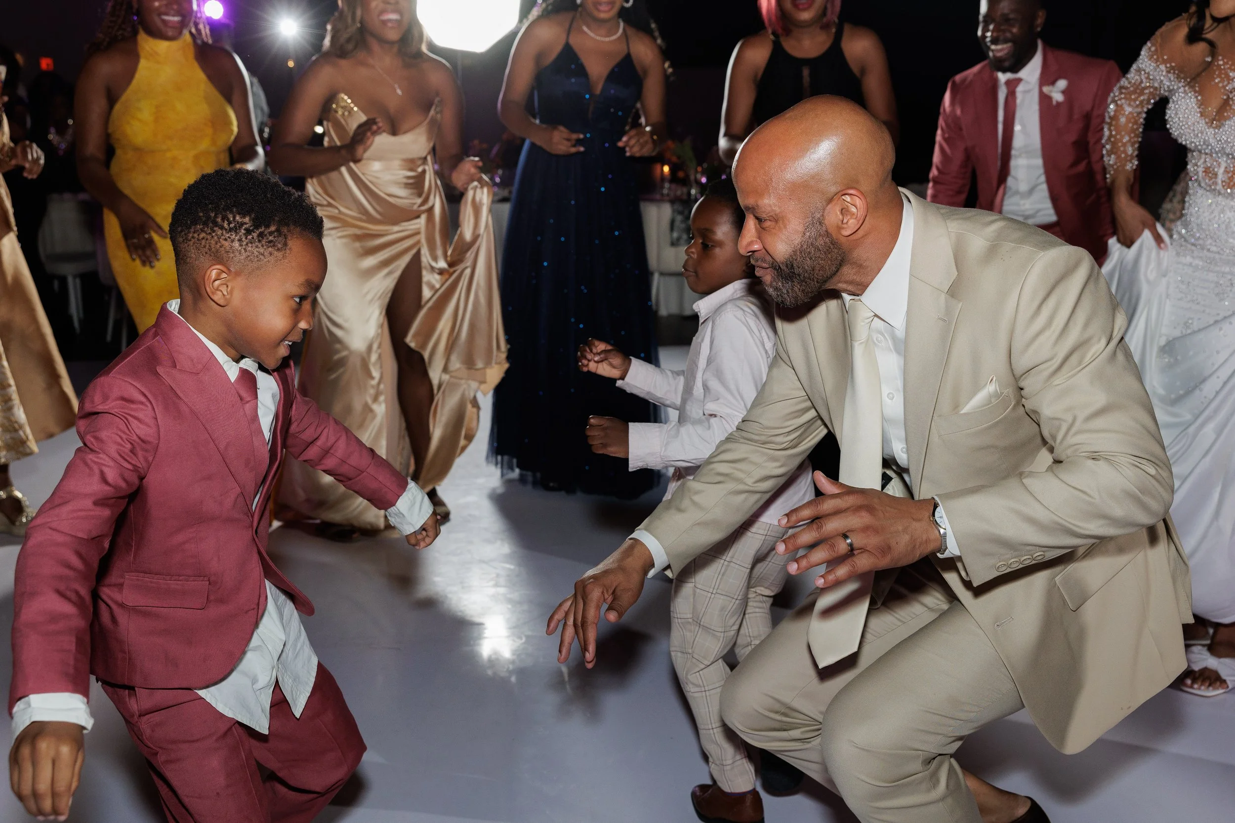 Man dancing with children at a celebration or wedding reception, with women in elegant dresses in the background.