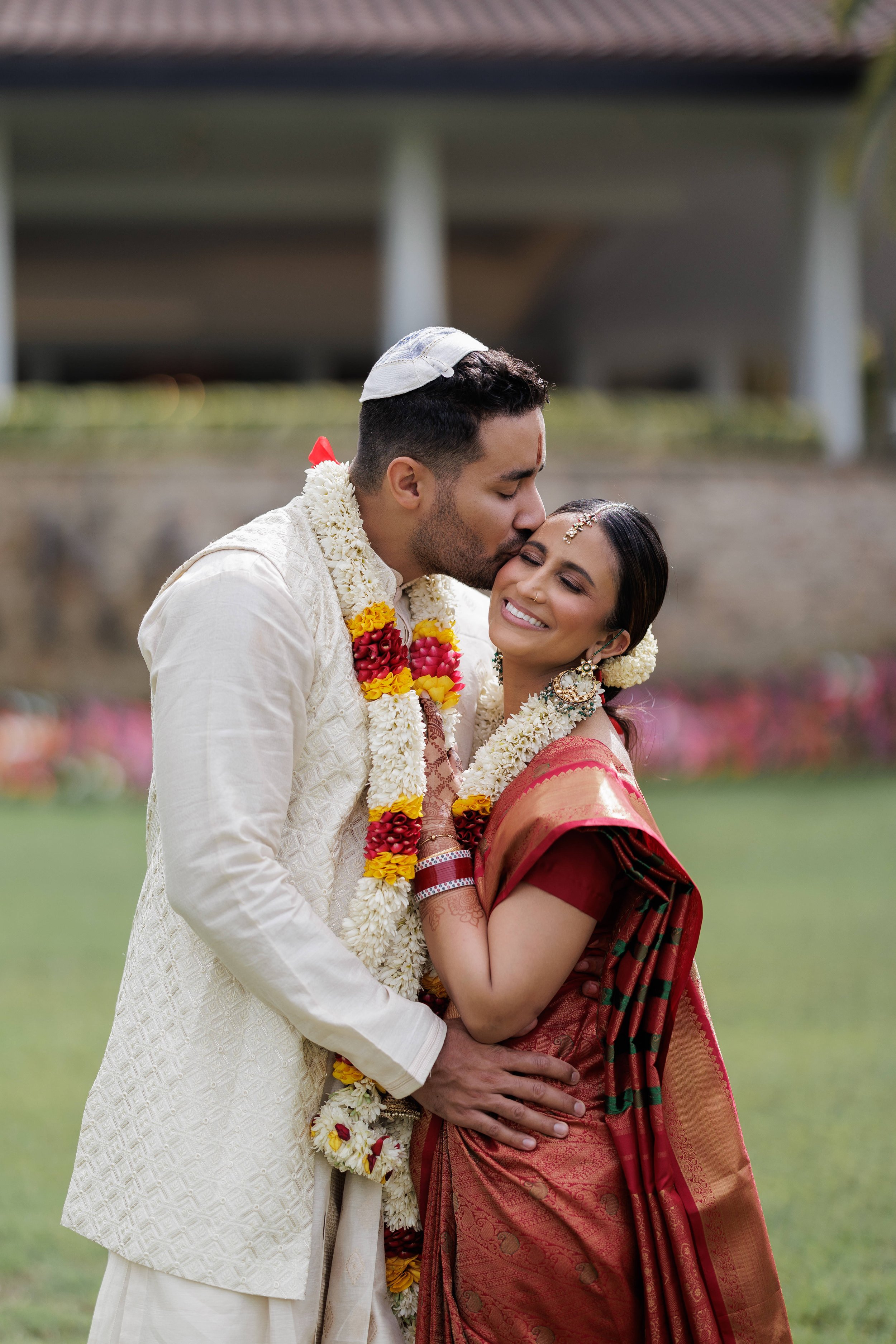 A newlywed couple in traditional Indian attire sharing a moment, with the groom giving a gentle kiss on the bride's forehead, both wearing flower garlands, outdoors in a garden setting.