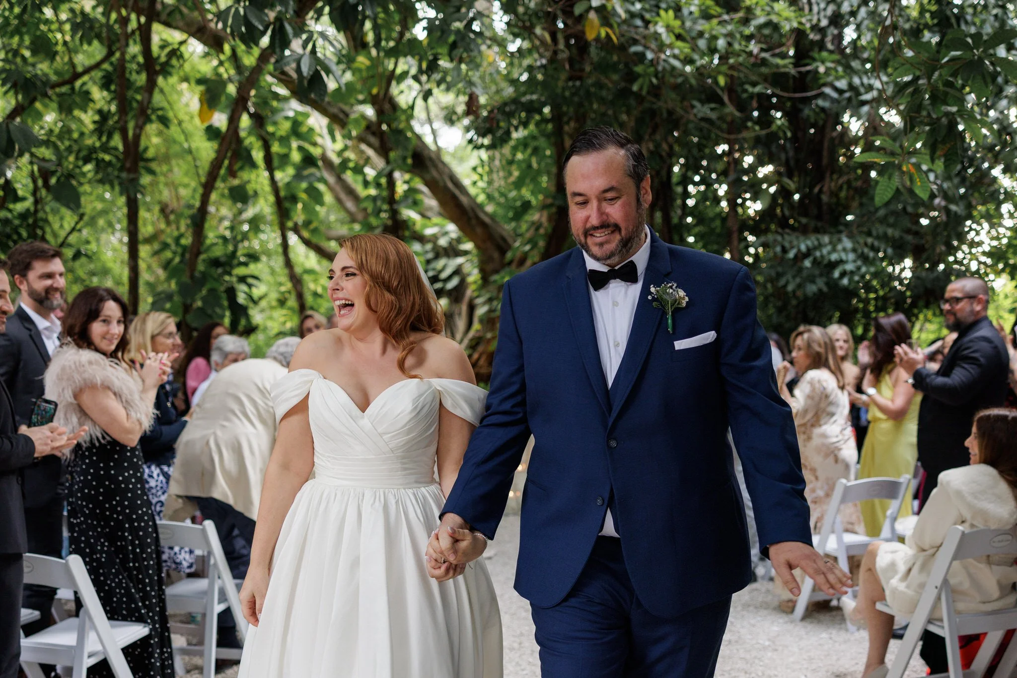 A bride and groom holding hands during their wedding, with the groom kissing the bride's forehead and the bride smiling, surrounded by tropical plants.