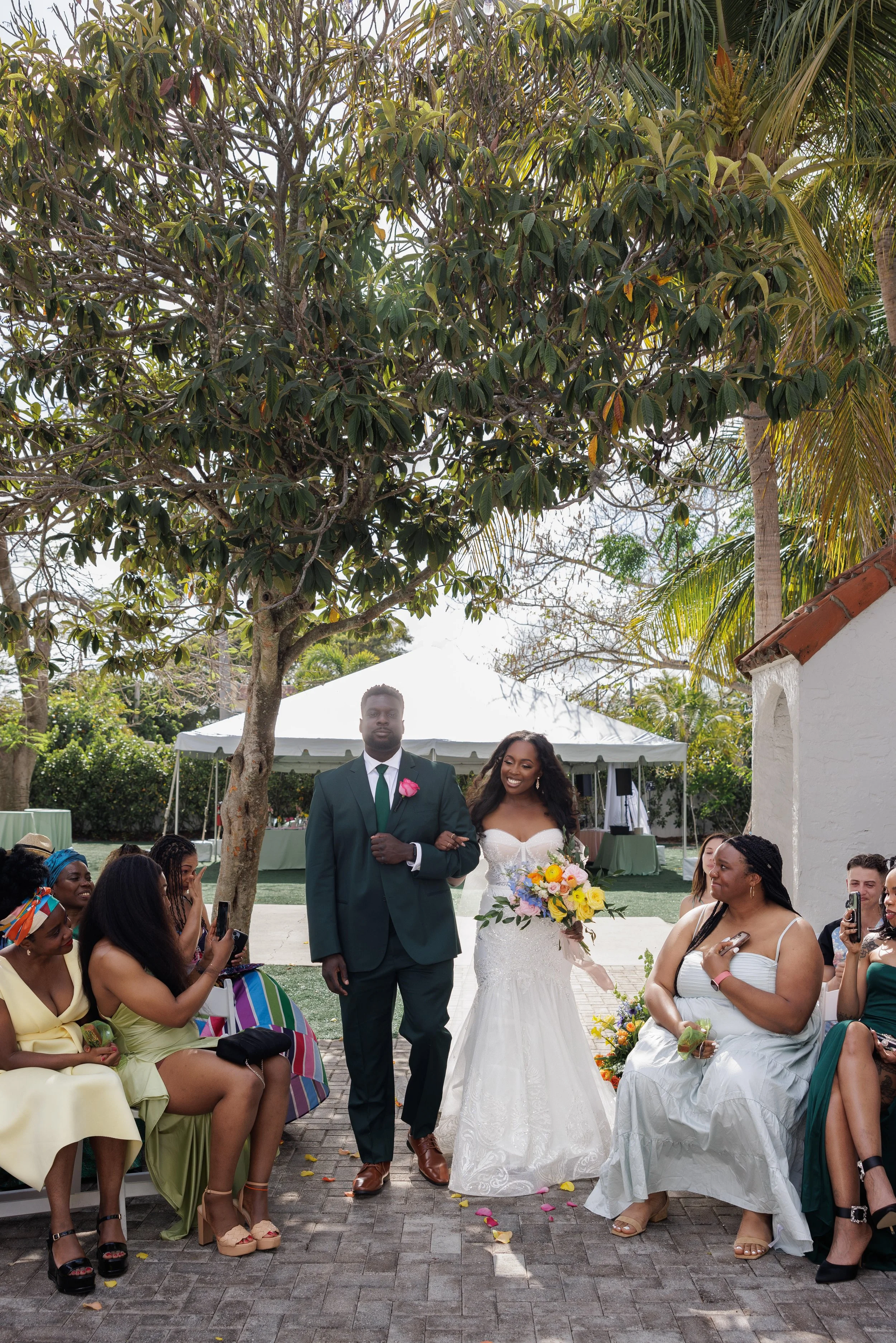 A wedding ceremony outdoors with a bride in a white lace gown holding a colorful bouquet, and a groom in a dark green suit with a pink boutonnière, walking arm in arm under a large tree. Guests are seated around, some taking photos, on a paved area s