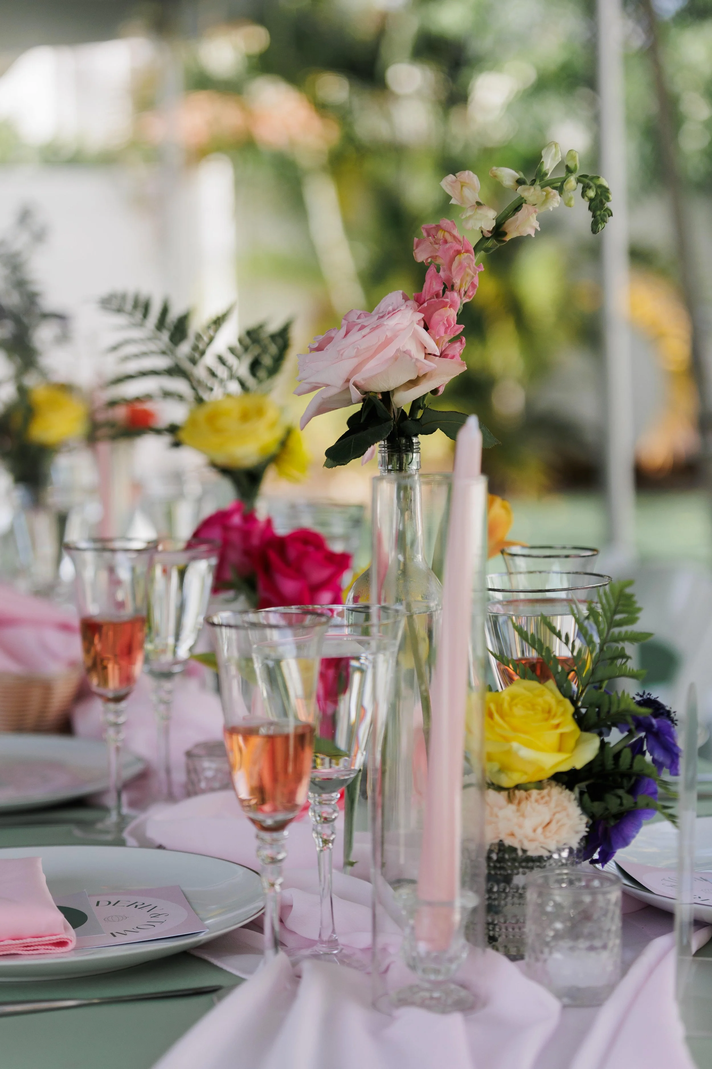 A table decorated with pink and yellow flowers in clear glass vases, pink candles, empty champagne glasses filled with rosé, and place settings with pink napkins and white plates, set outdoors with blurred greenery in the background.
