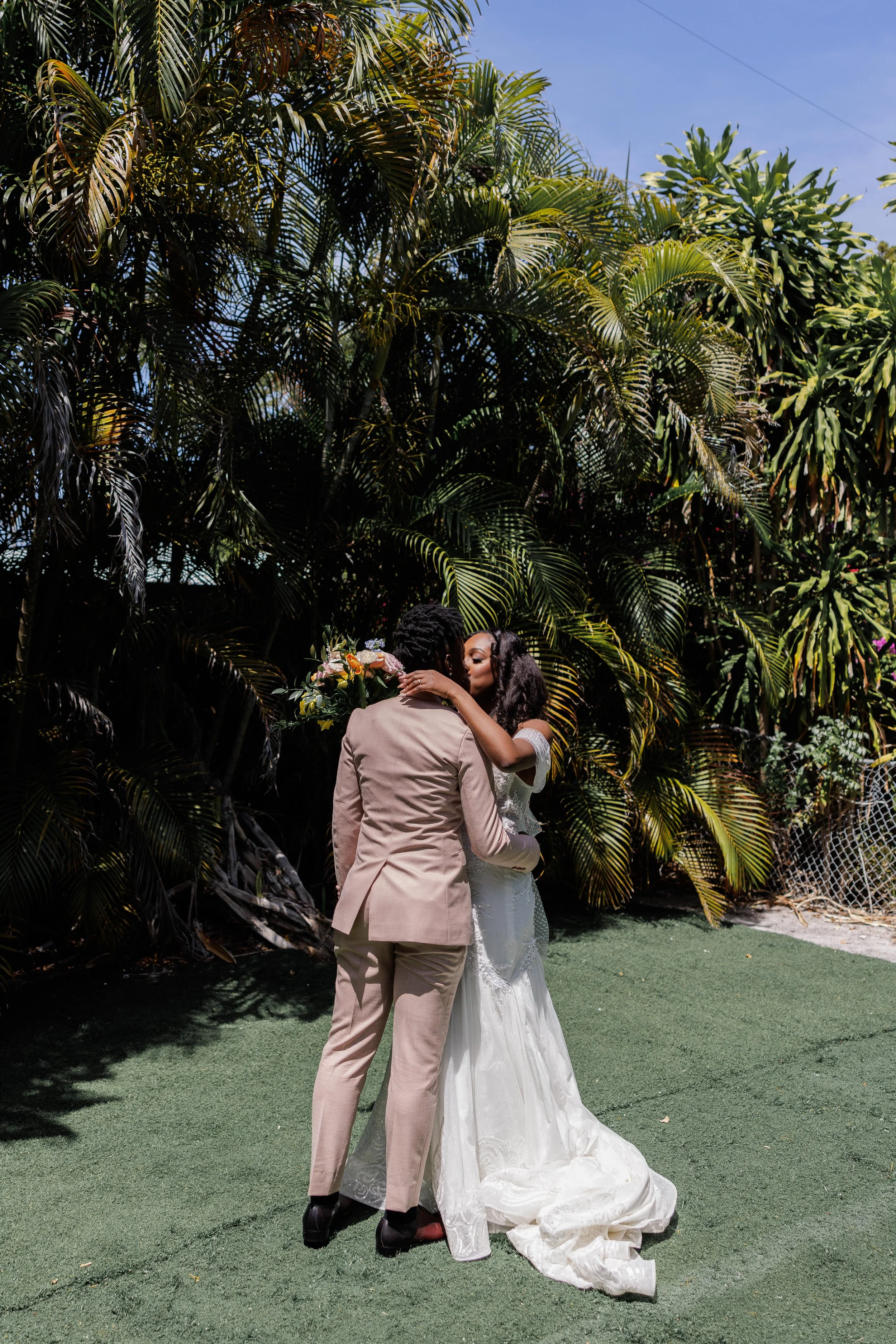 A couple dressed in wedding attire sharing a kiss in a lush garden with large green tropical plants.
