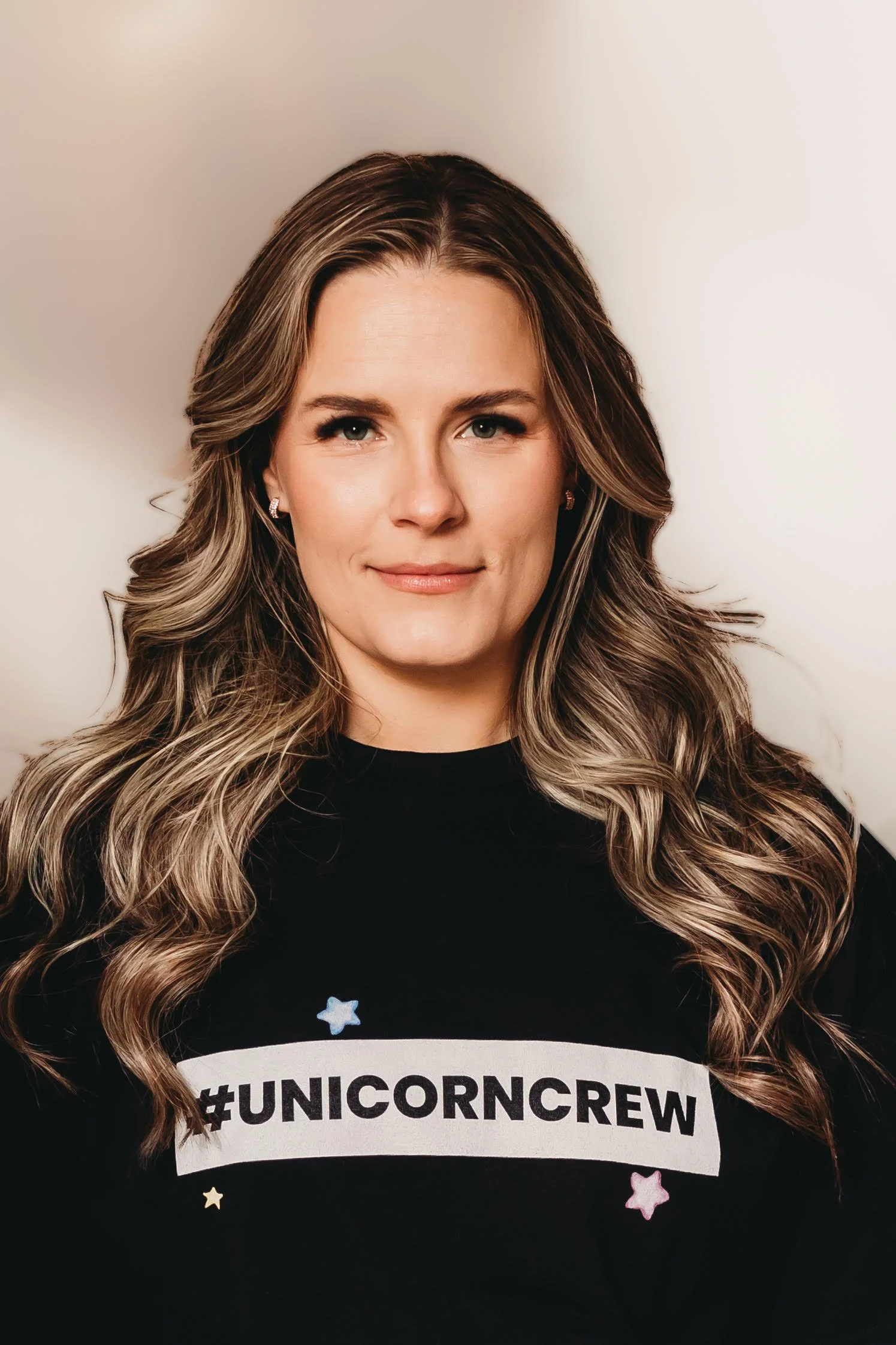 A woman with long, wavy, light brown hair wearing a black shirt with the hashtag #UNICORNCREW and small colorful star designs.