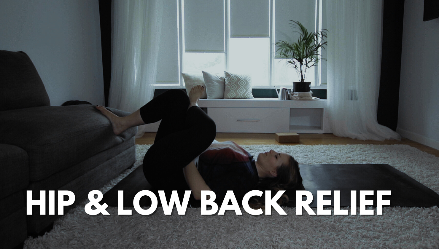 A woman lying on her back on a yoga mat doing a hip and low back stretch in a living room. There is a sofa, a window with curtains, a potted plant, and a white cabinet in the background.