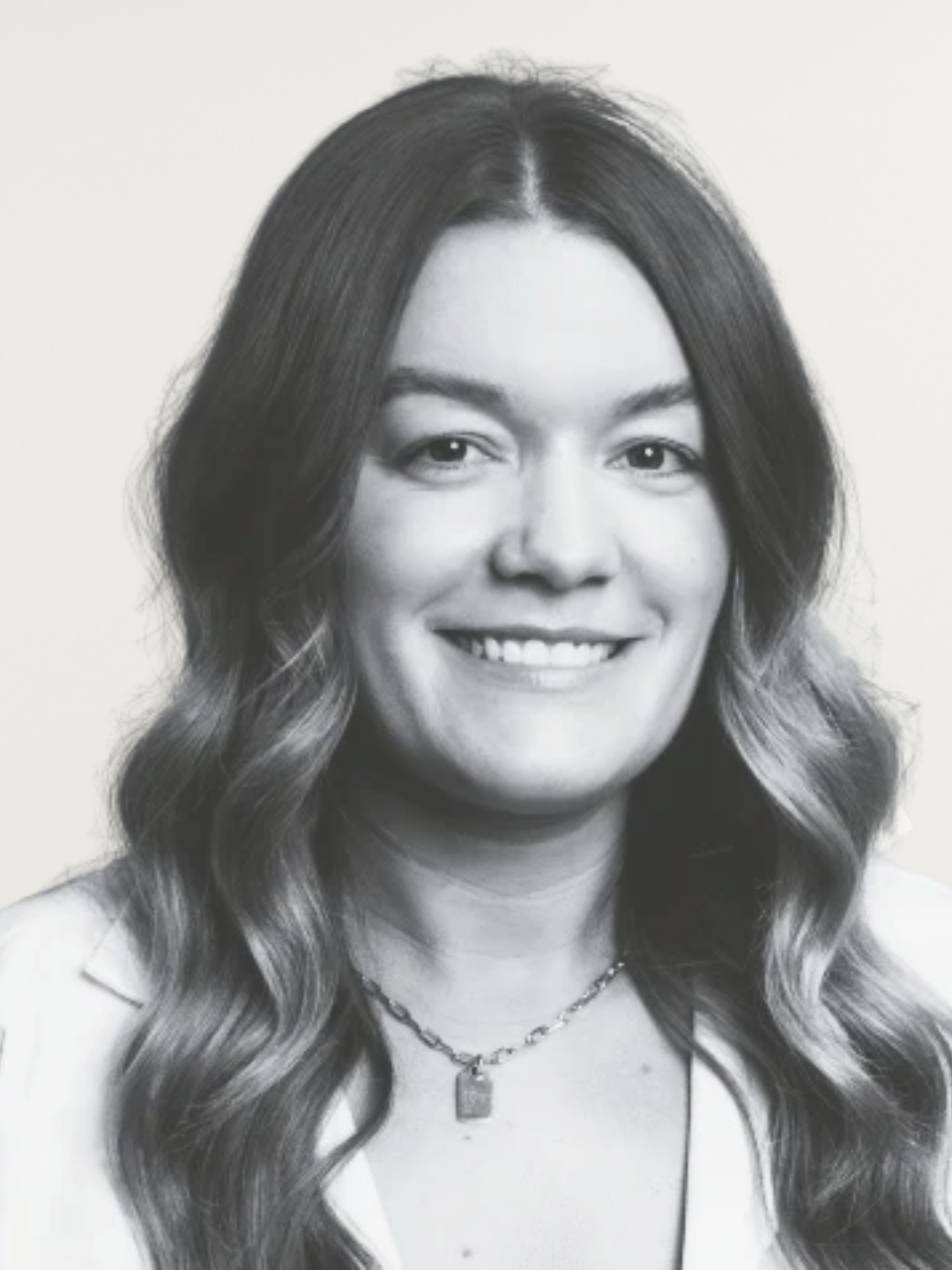 Black and white portrait of a young woman with long wavy hair, smiling, wearing a light-colored shirt and a necklace, against a plain background.
