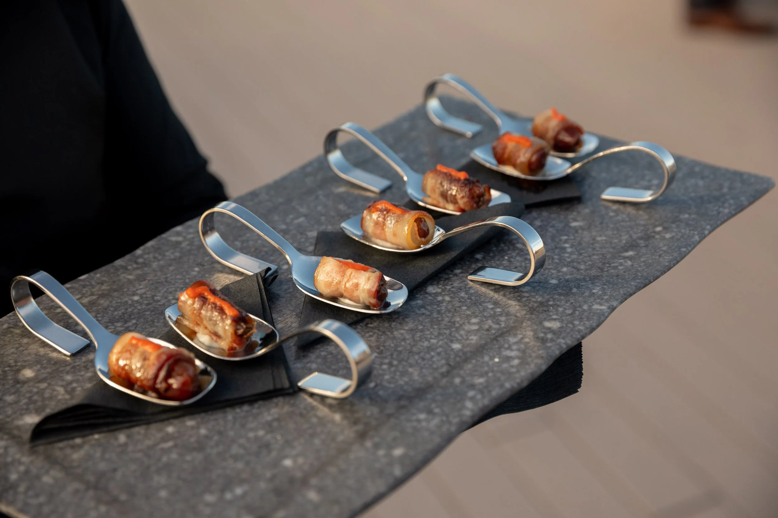 Bacon wrapped dates served on large silver spoons on a marble tray.