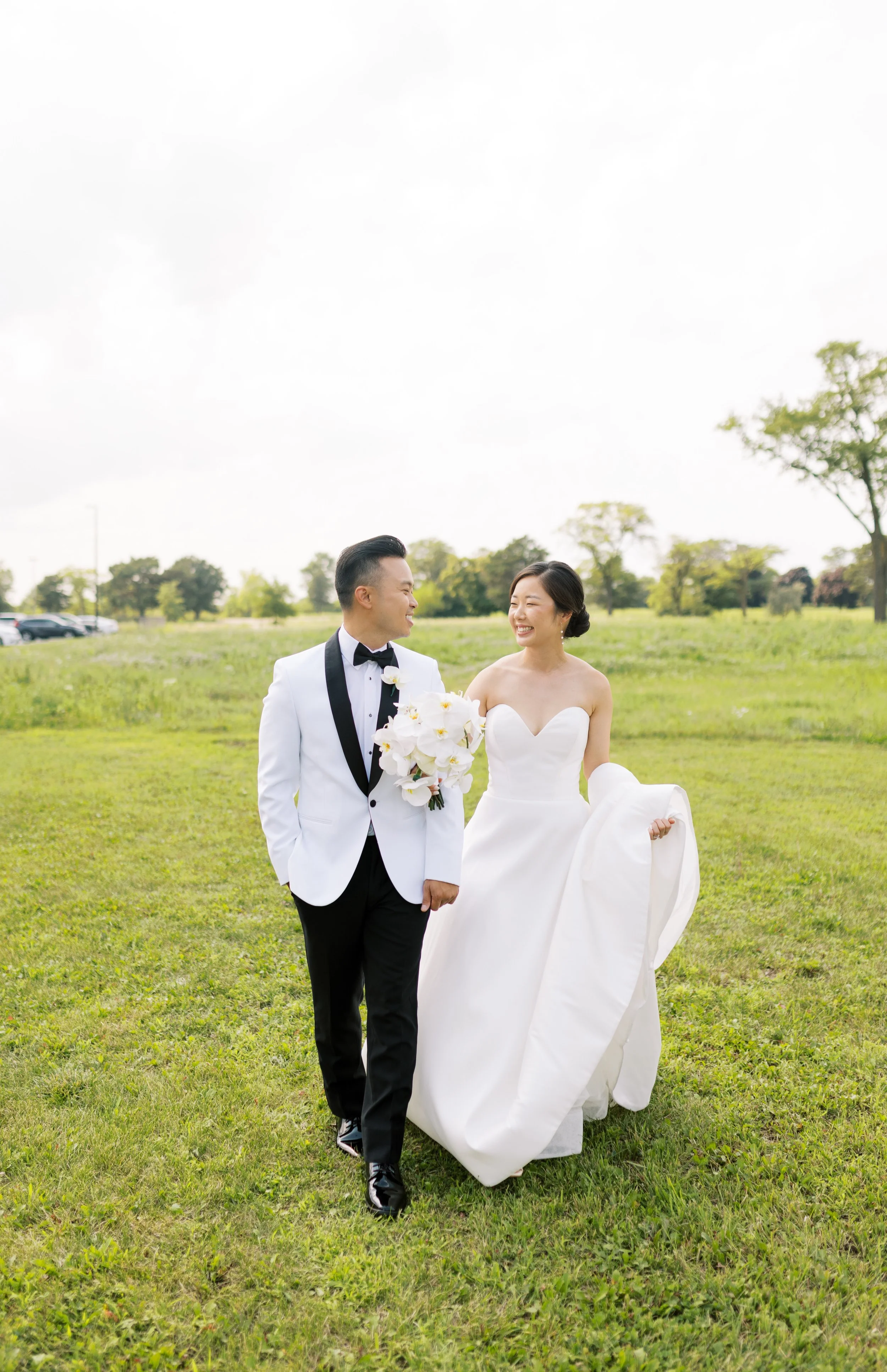 A newlywed couple walking and smiling in a grassy outdoor setting during daytime, the groom in a white tuxedo and the bride in a strapless white wedding gown holding a bouquet of white flowers.