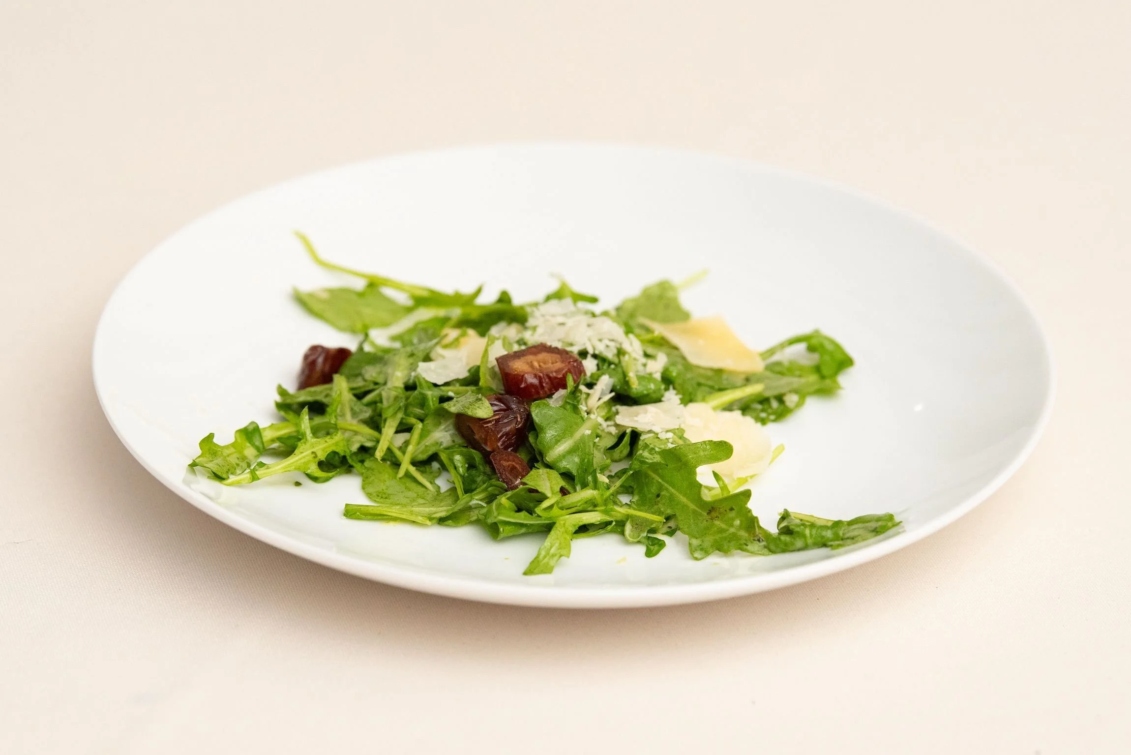 Arugula salad served on a round white plate with parmesan, dates, beets, and dressing.