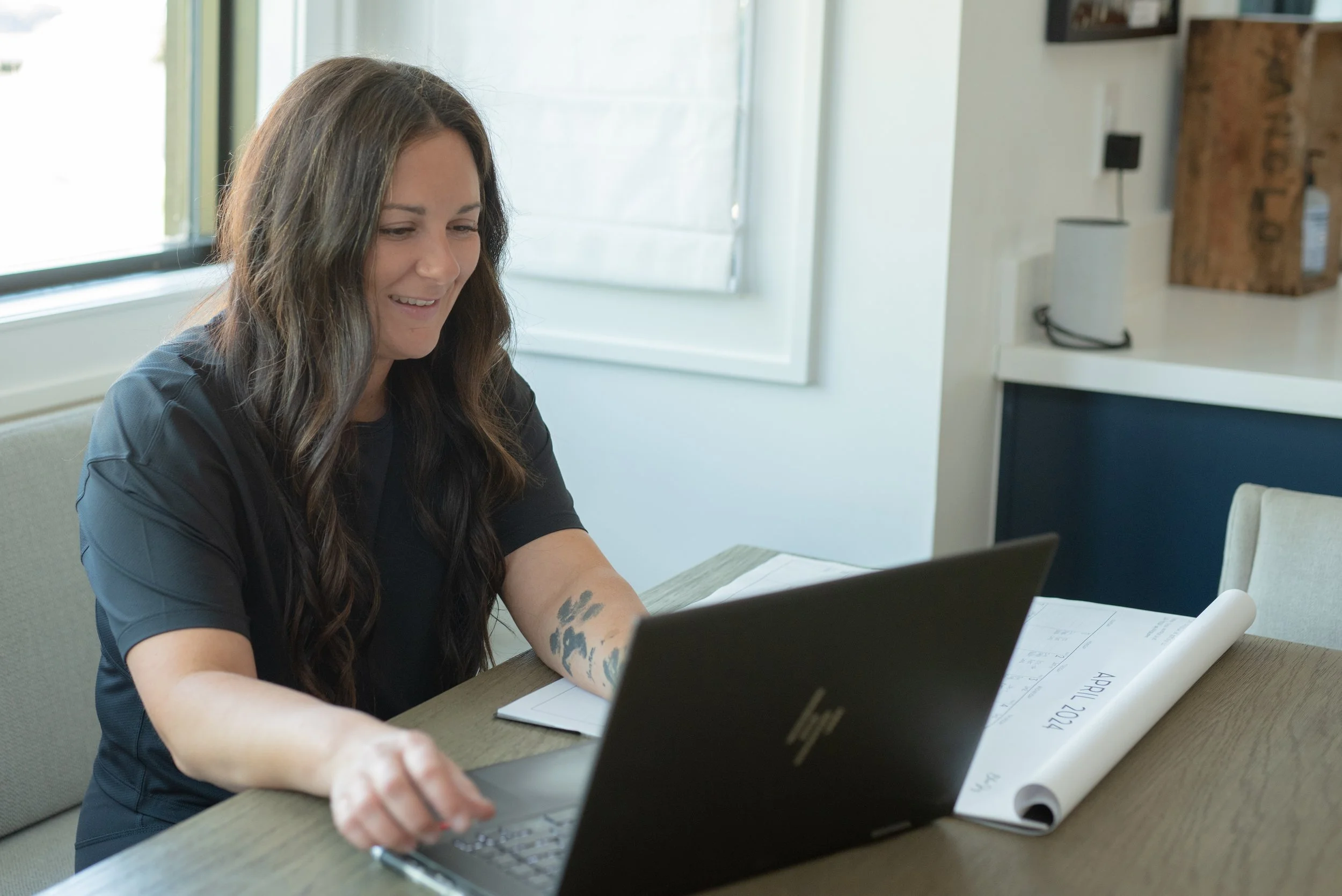 A woman with long brown hair sitting at a table, working on a laptop, with a calendar on the table showing April 2024.