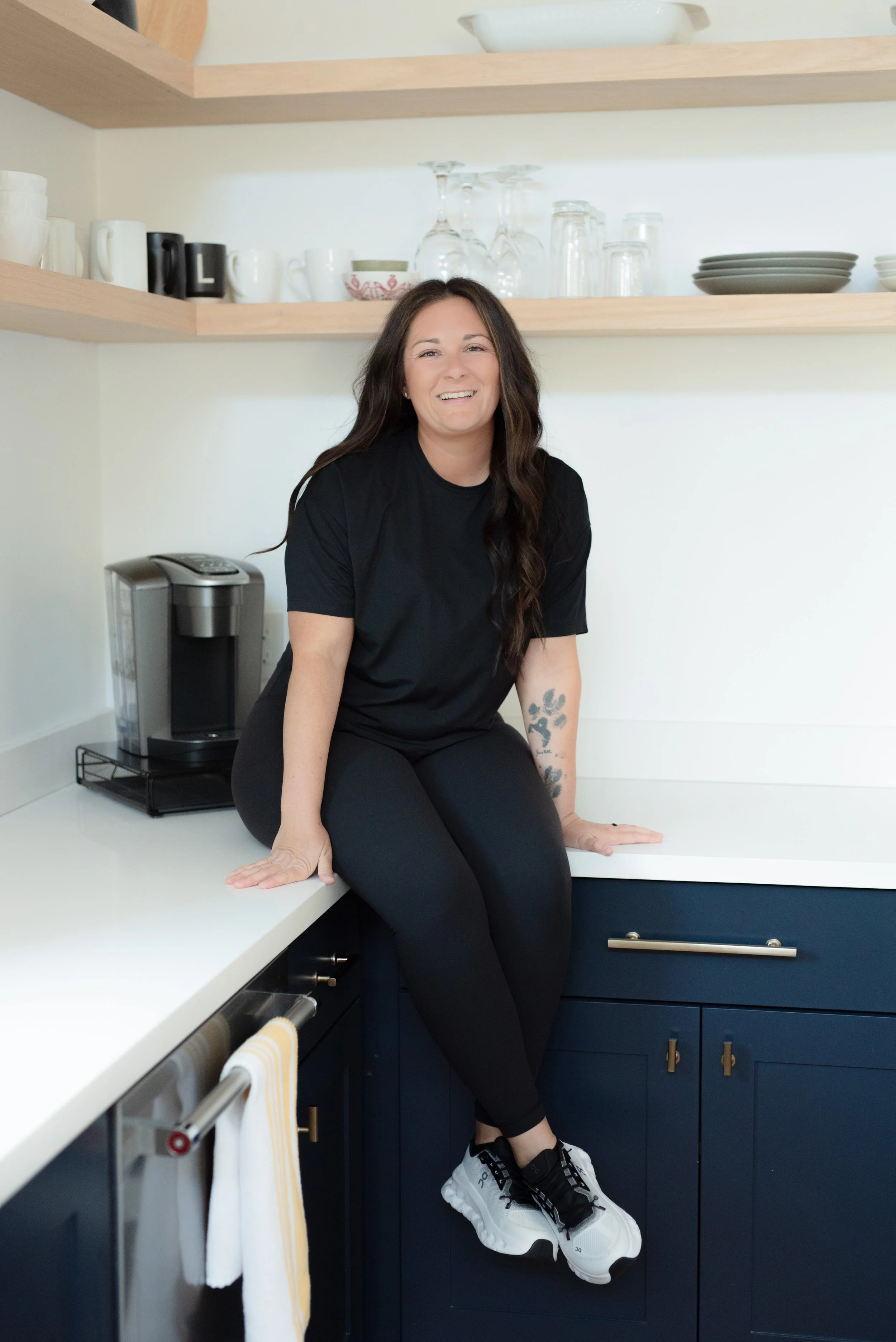 A woman with long dark hair, wearing a black t-shirt and black pants, sits on a kitchen counter smiling. The kitchen has open wooden shelves with dishes and glassware, and a coffee maker on the counter.