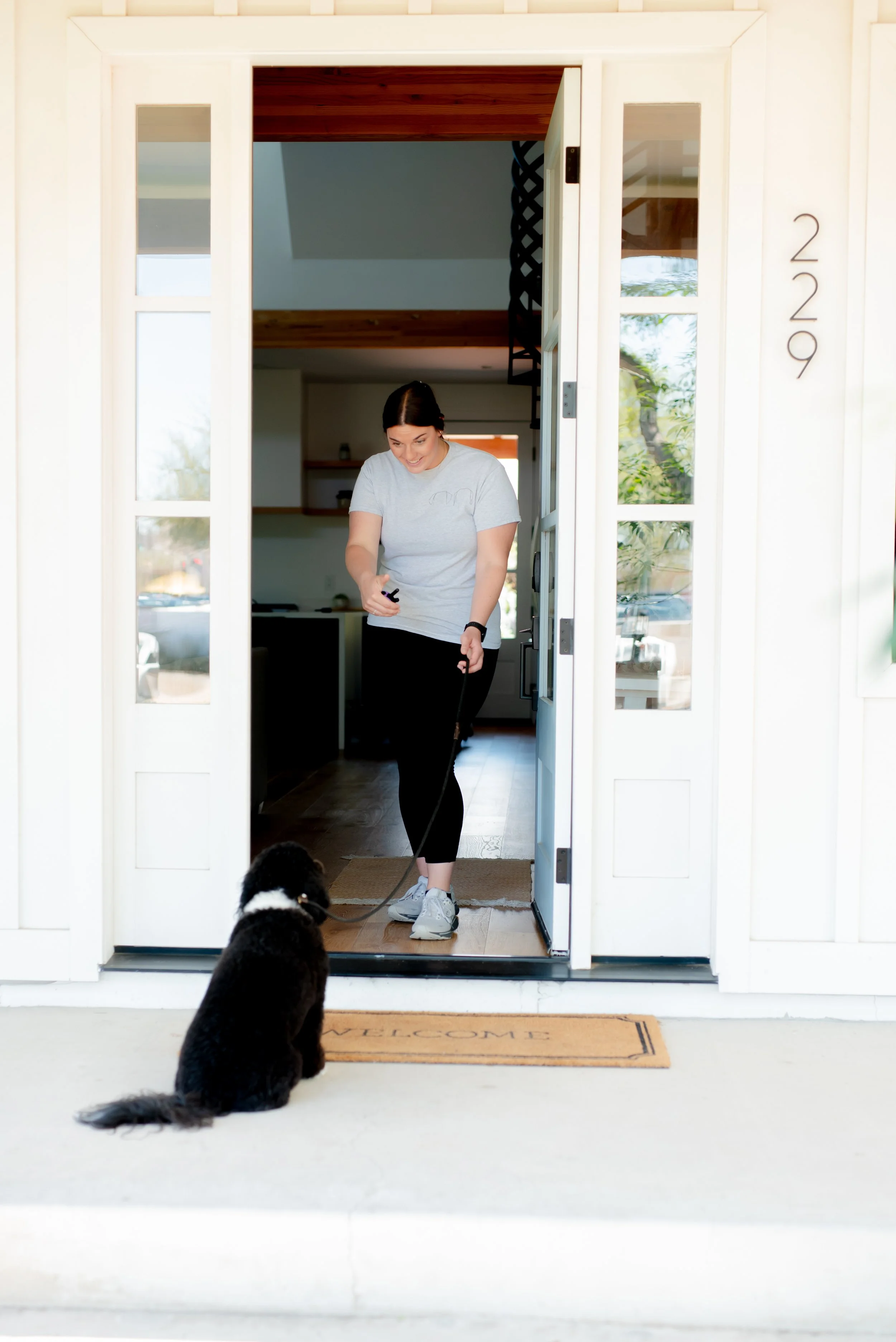 A woman standing at her front door with a black and white dog sitting on the welcome mat outside, smiling while looking at her phone.