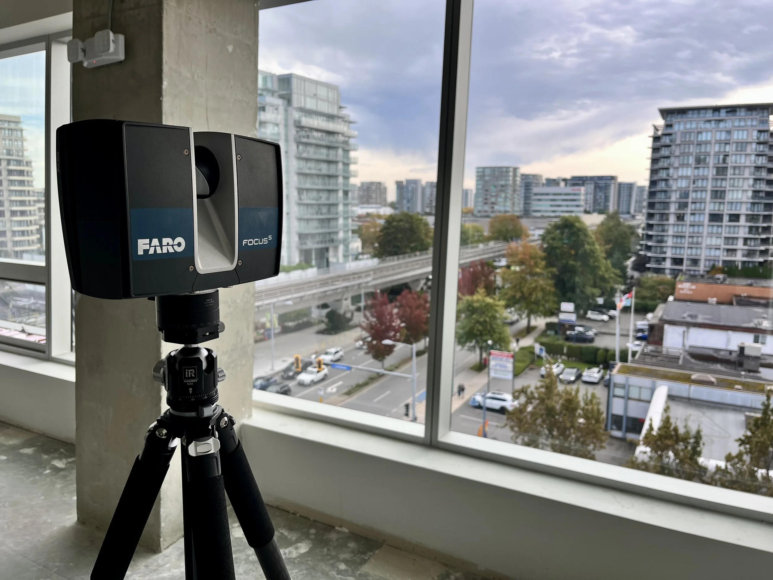 Lidar sensor on a tripod inside a building with large windows overlooking a cityscape with residential buildings and trees.