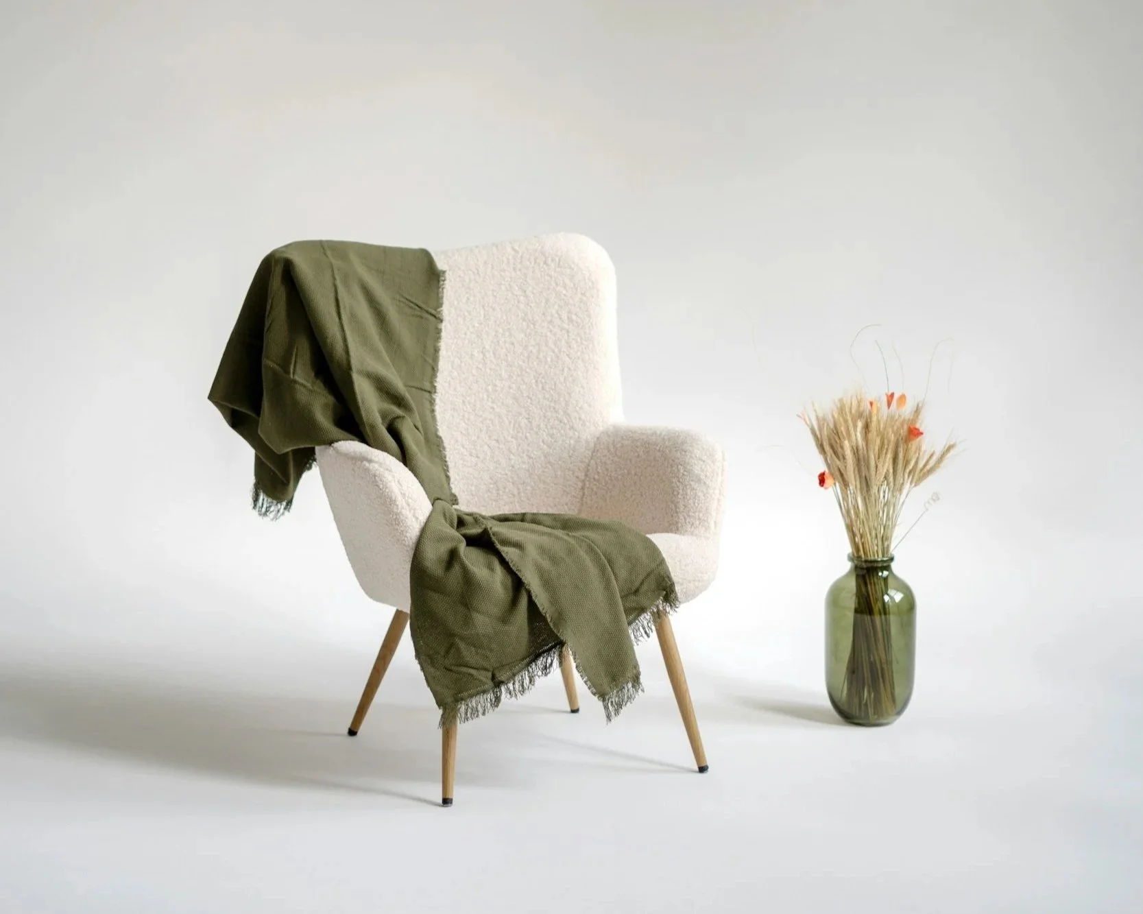 A white upholstered armchair with wooden legs, draped with a green fabric blanket, positioned next to a tall green glass vase filled with dried flowers.