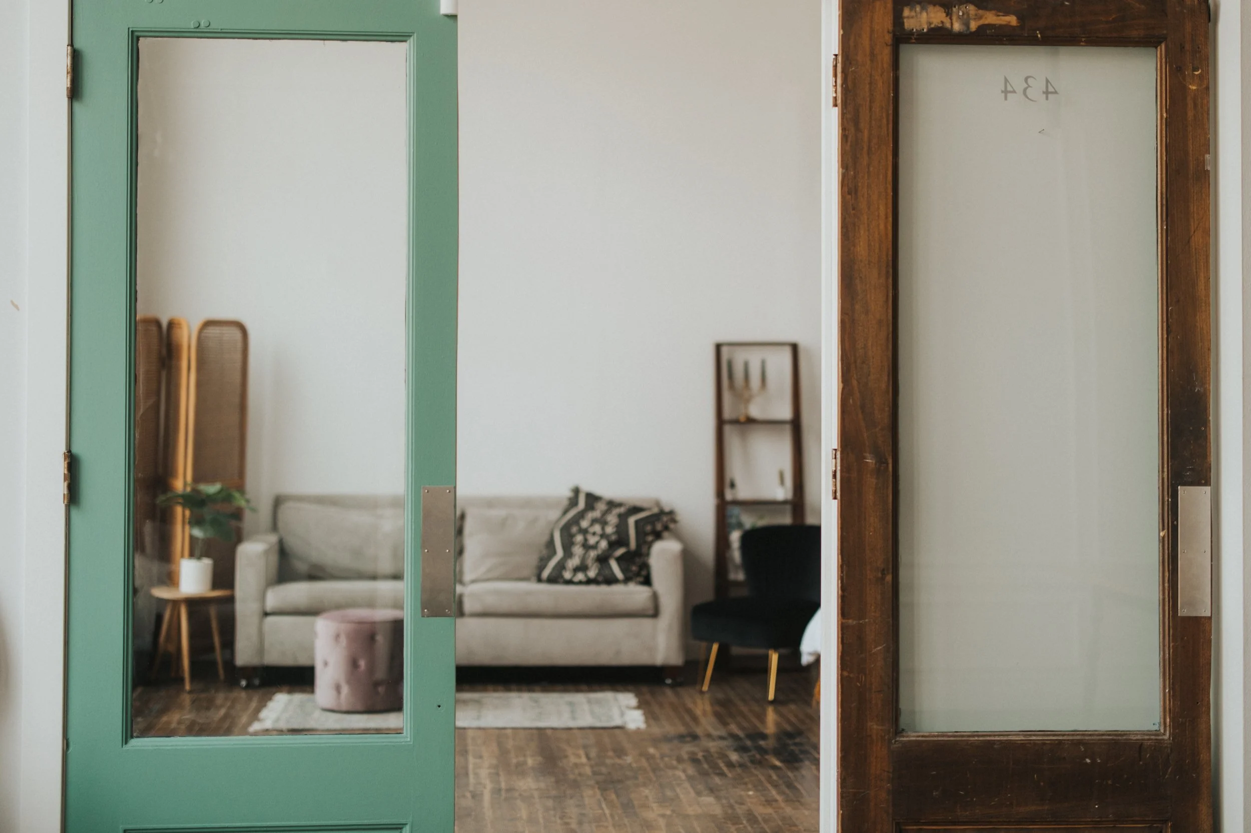Living room seen through two glass-paneled doors, one green and one wooden, both slightly open. The room has a white couch with black and white pillows, a small round pink ottoman, a wooden side table with a lamp and plant, and a framed artwork on the wall.