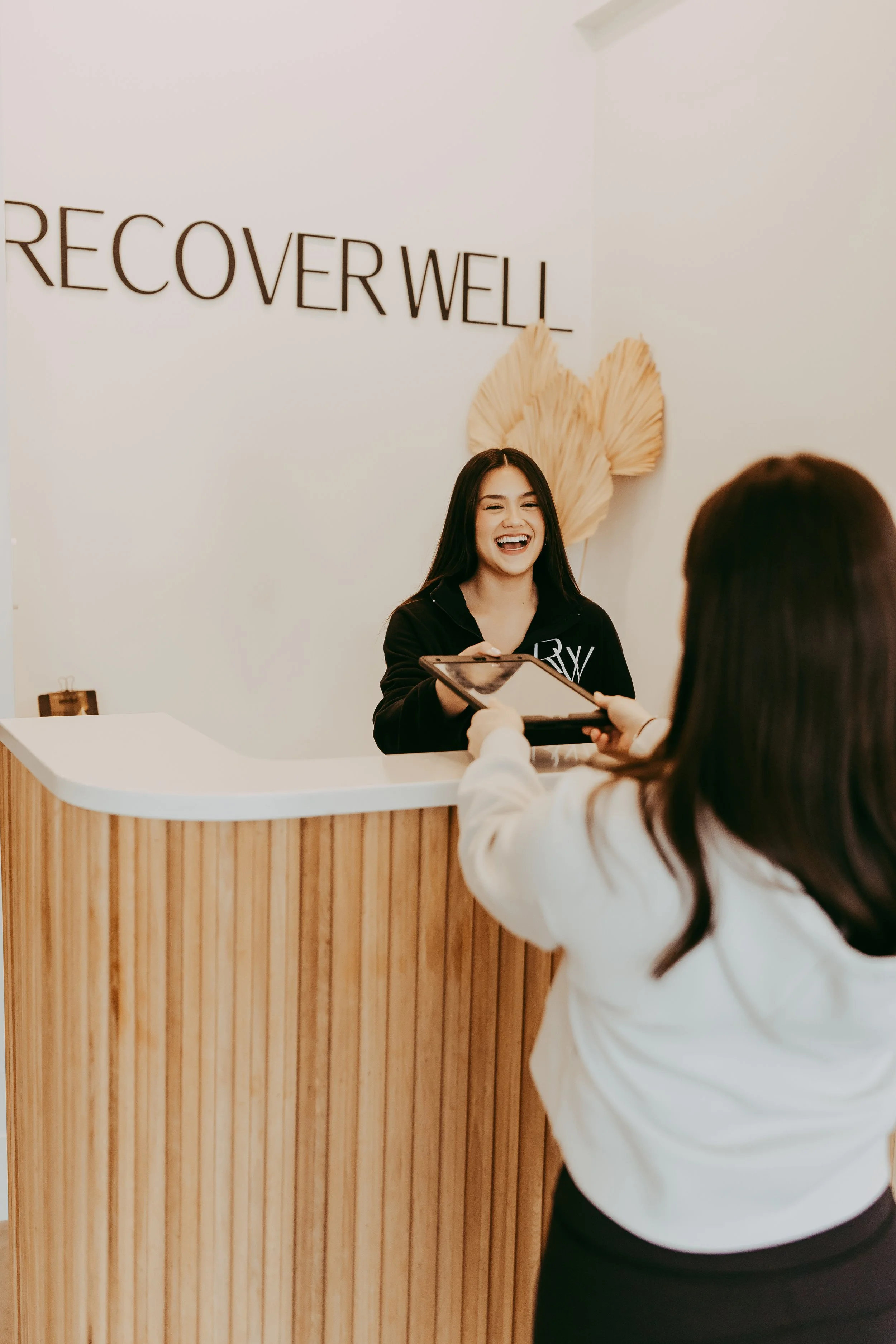 Two women, one dressed in a white robe with fuzzy earmuffs and gloves, smiling and walking out of a cryotherapy chamber, the other woman in casual clothing smiling and looking towards her.