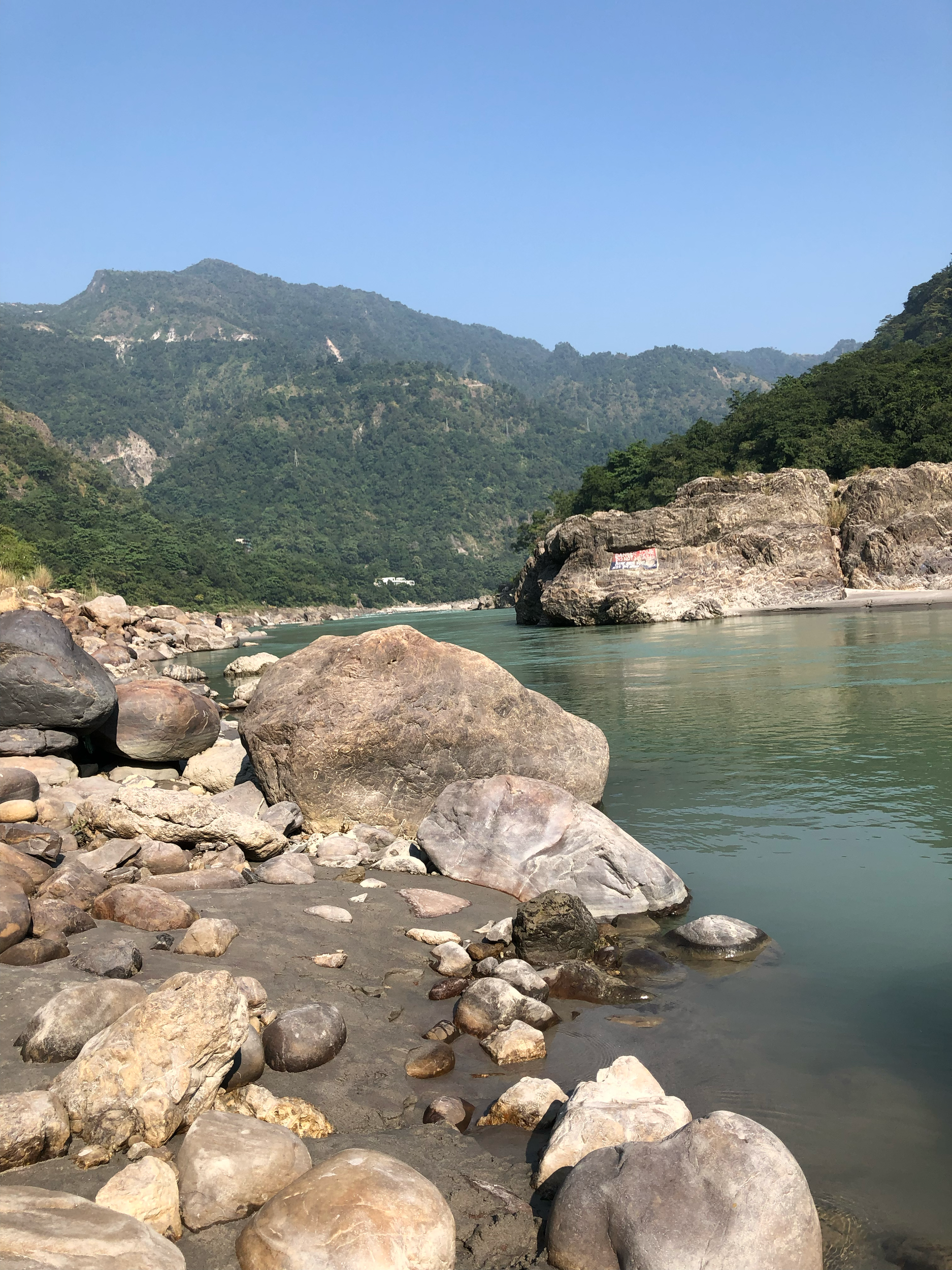 Rocks along the shore of a river with mountains and greenery in the background under a clear blue sky.