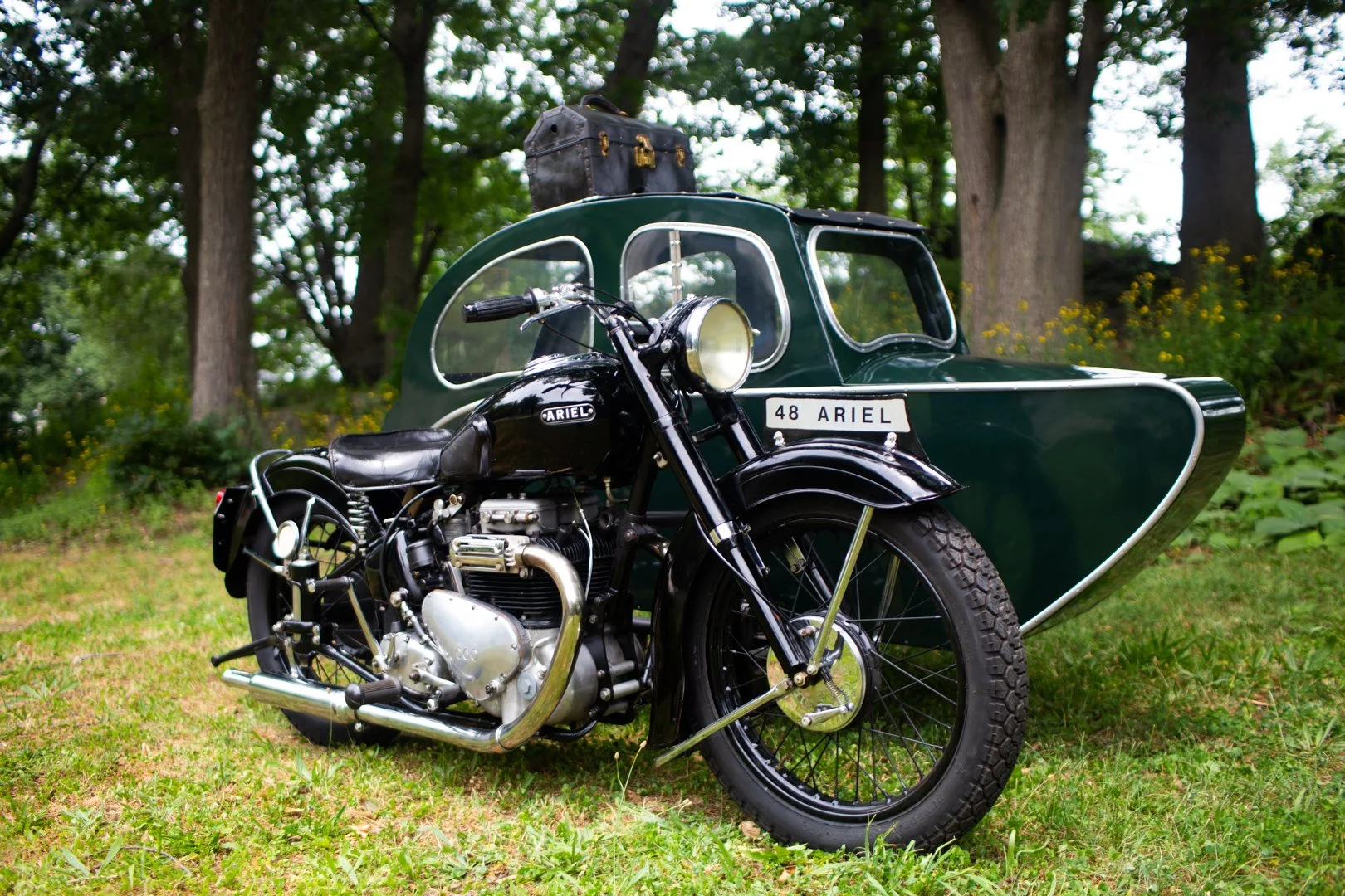A vintage black Ariel motorcycle with a sidecar parked on grass, surrounded by trees.