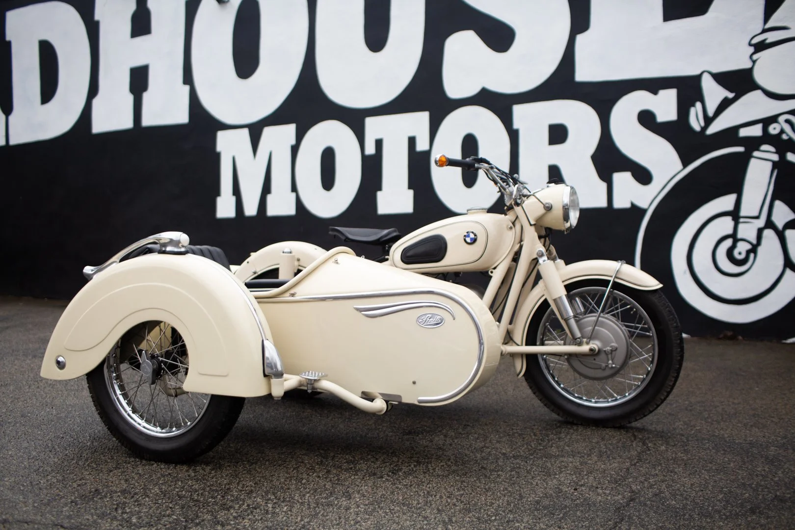 Vintage cream-colored BMW motorcycle with sidecar parked on a paved surface in front of a black wall with white text and illustrations of motorcycles.