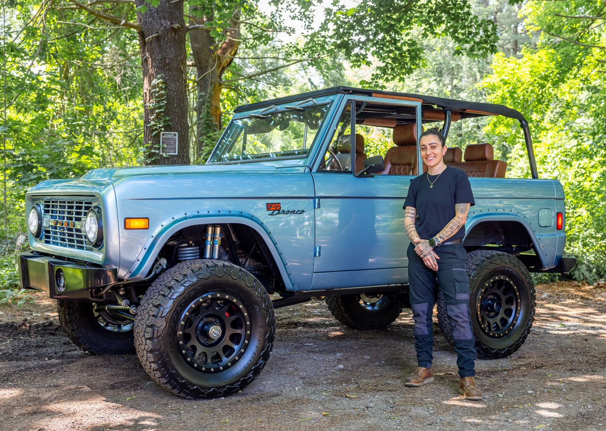 A woman standing next to a vintage blue Ford Bronco with brown leather seats in a forest setting.