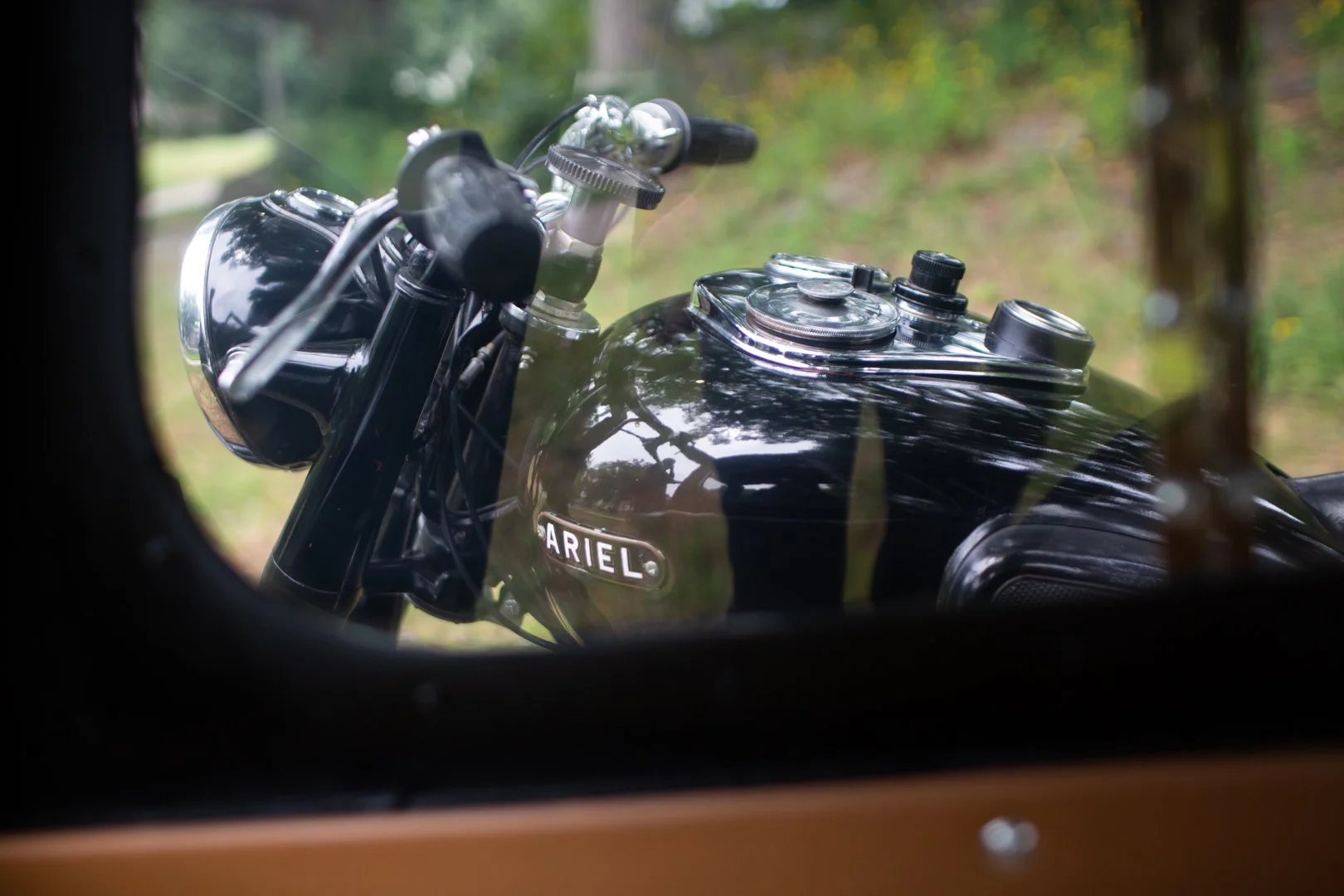 Close-up view of a black Ariel motorcycle tank seen through a small window or opening, with reflections of trees and sky, and a blurred background of green foliage.