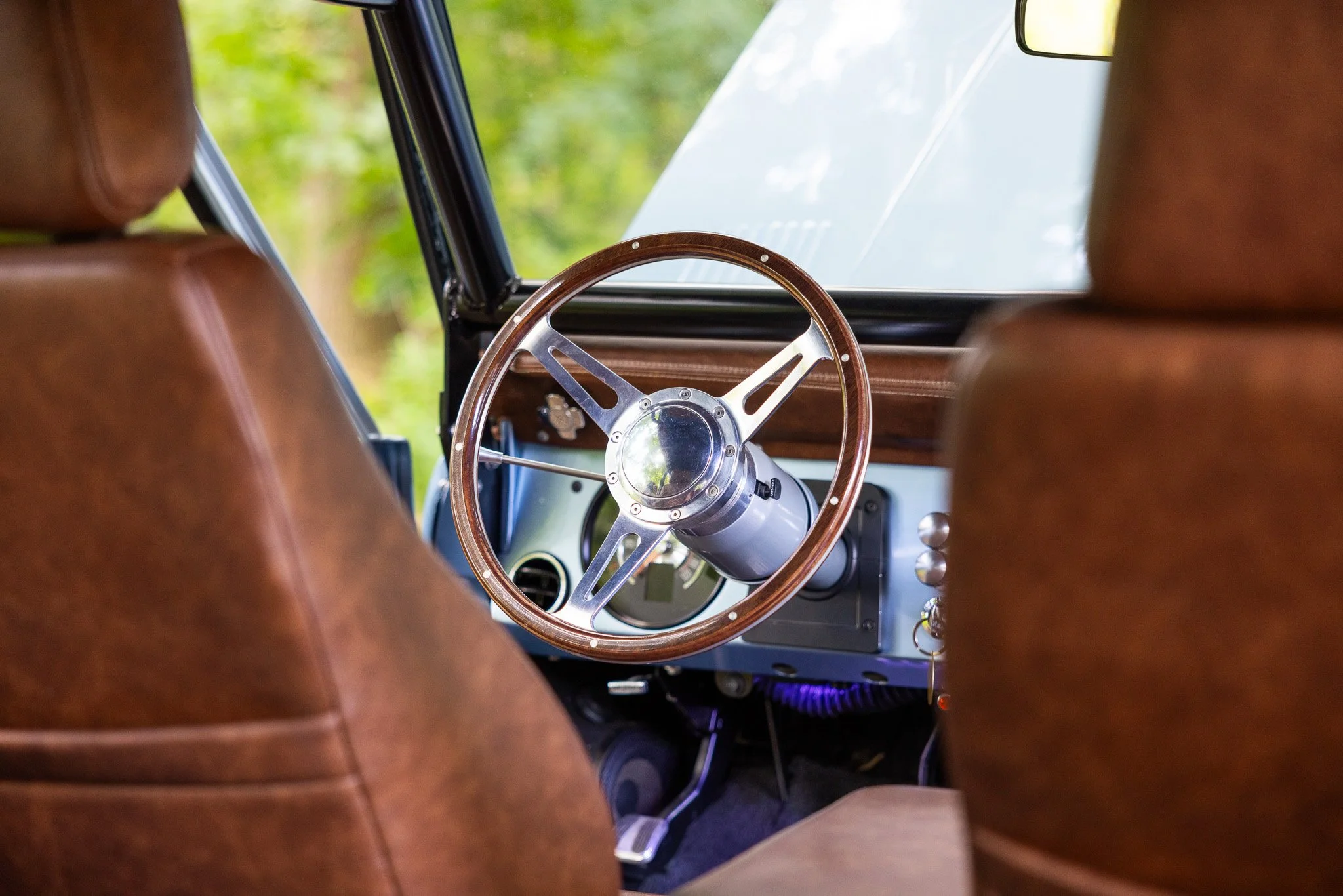 View of a car interior from the backseat, showing a vintage-style wooden steering wheel, dashboard, and front seats with brown upholstery. Through the windshield, a blurred view of green trees is visible.