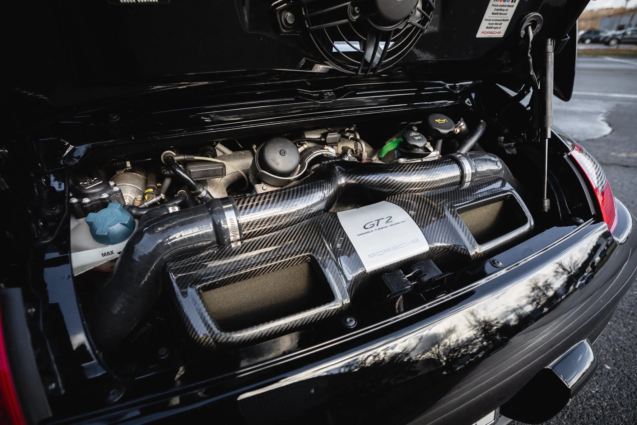 Close-up of a Porsche Carrera GT engine, showing a carbon fiber intake and various engine components, with the car's hood open in a parking lot.