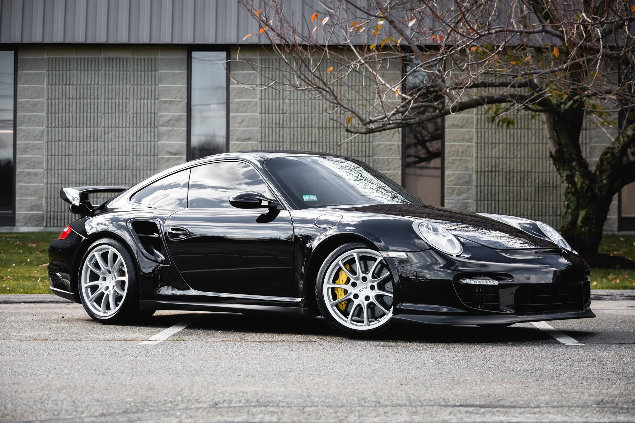 Black Porsche 911 sports car parked in an outdoor parking lot with a building and tree in the background.