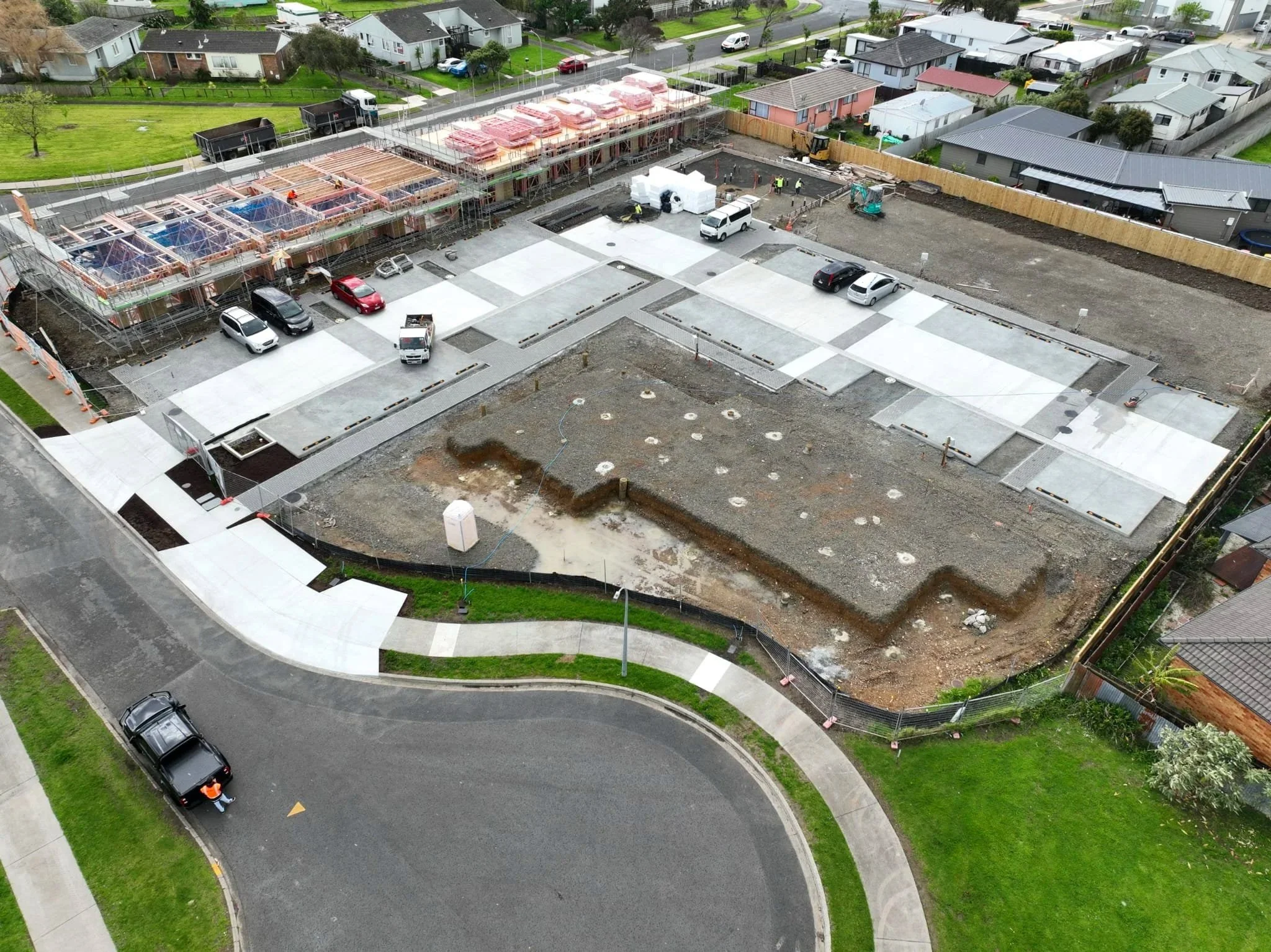 An aerial view of a construction site with several workers, parked cars, and construction materials, surrounded by residential houses and greenery.