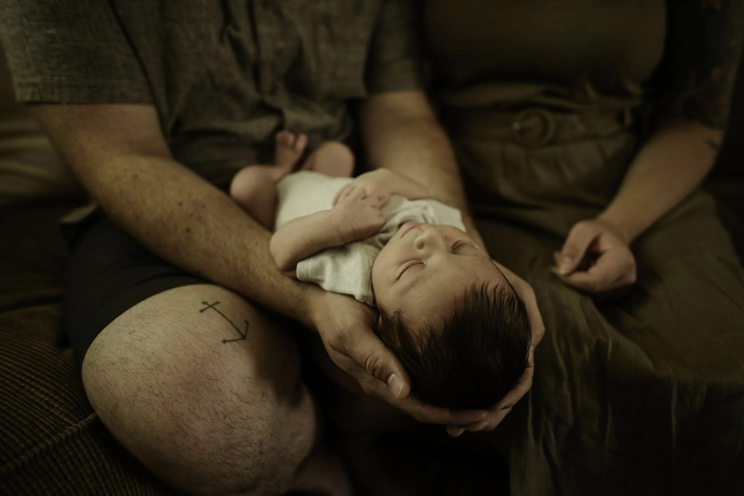 A newborn baby lying on an adult's lap, with the adult holding the baby's head and gently supporting their back. The baby's eyes are closed, and the scene appears to be in a cozy indoor setting.