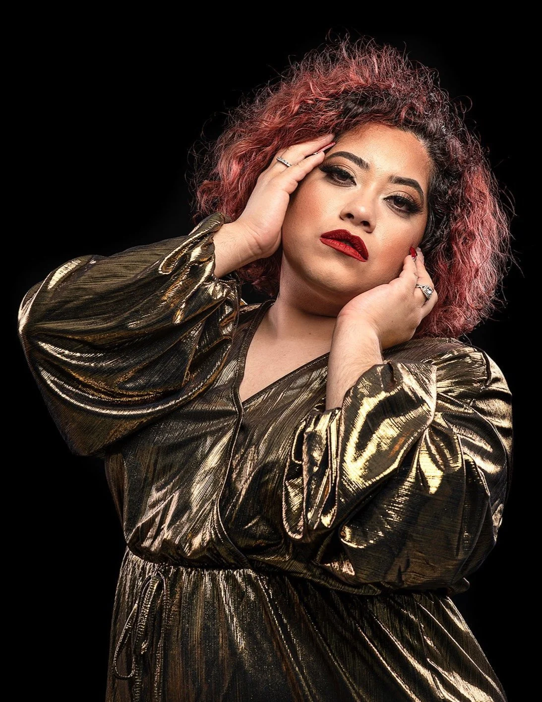 A woman with curly pink hair wearing a shiny metallic gold dress poses against a black background, with one hand on her face and the other touching her cheek.