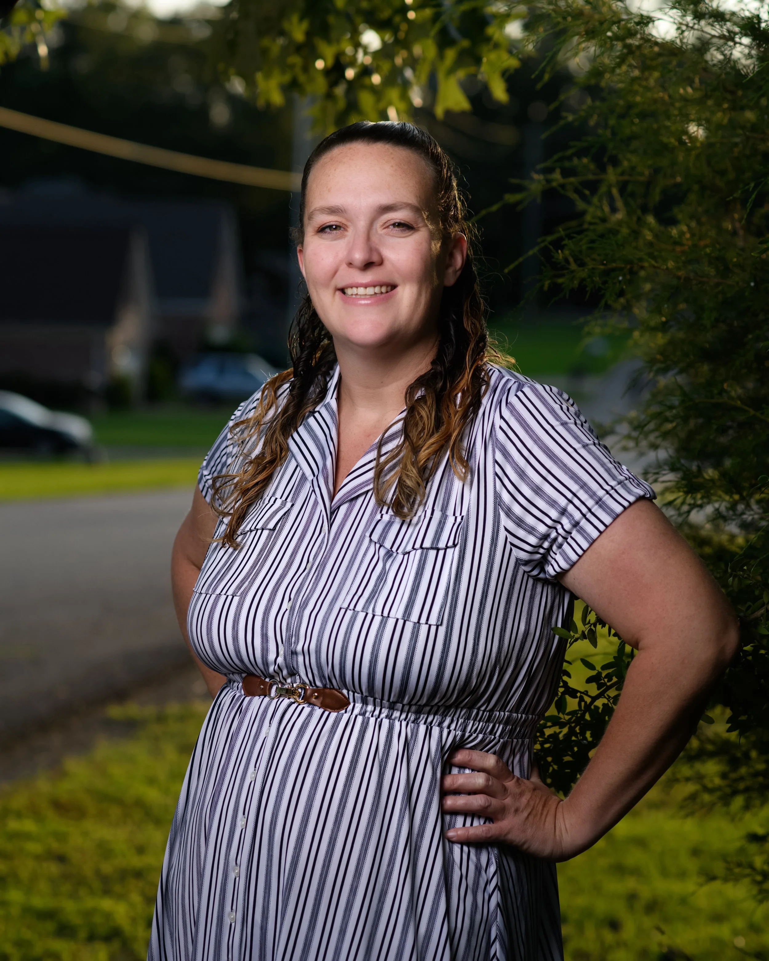 A woman standing outdoors near greenery, smiling, with her hand on her hip, wearing a striped dress with a brown belt.