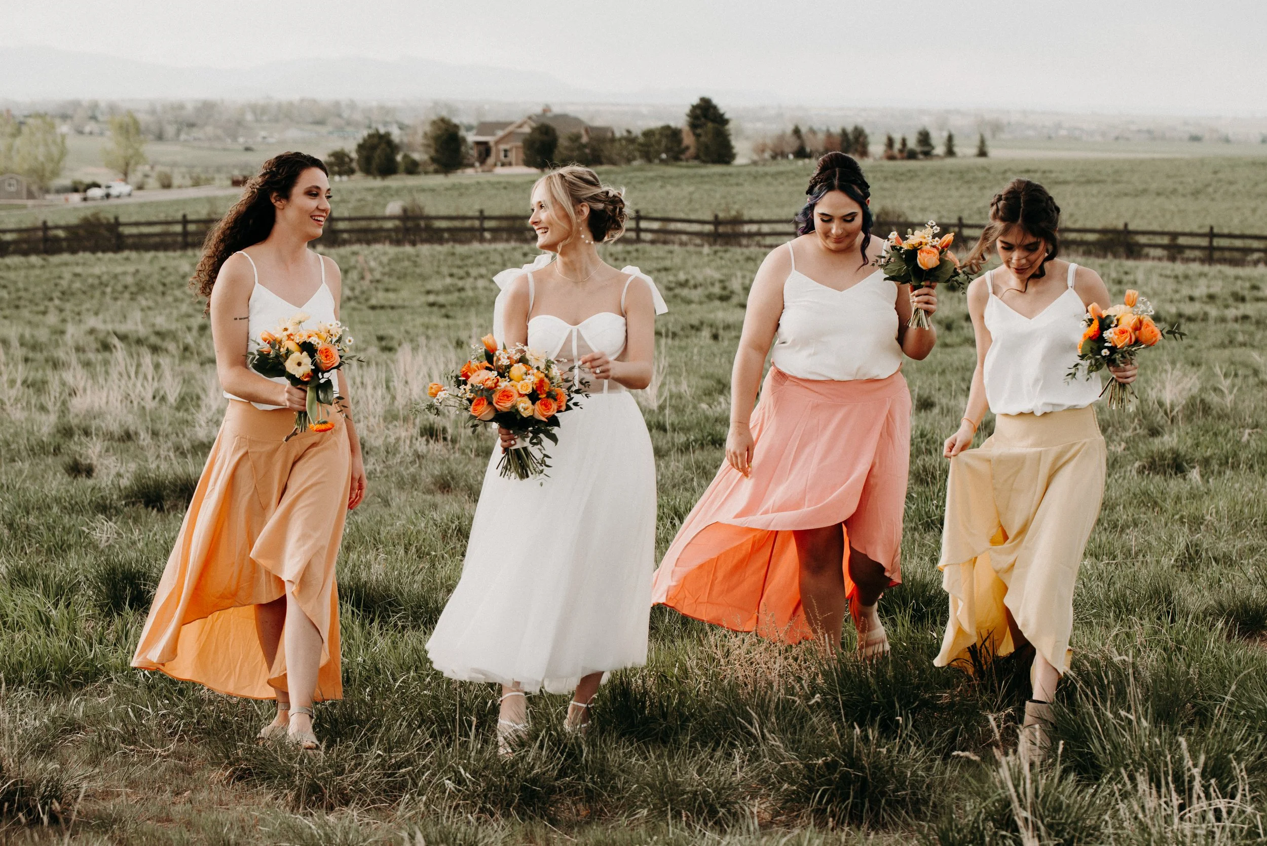 Five women in dresses walking through a field, holding bouquets of flowers, with a rural landscape in the background.