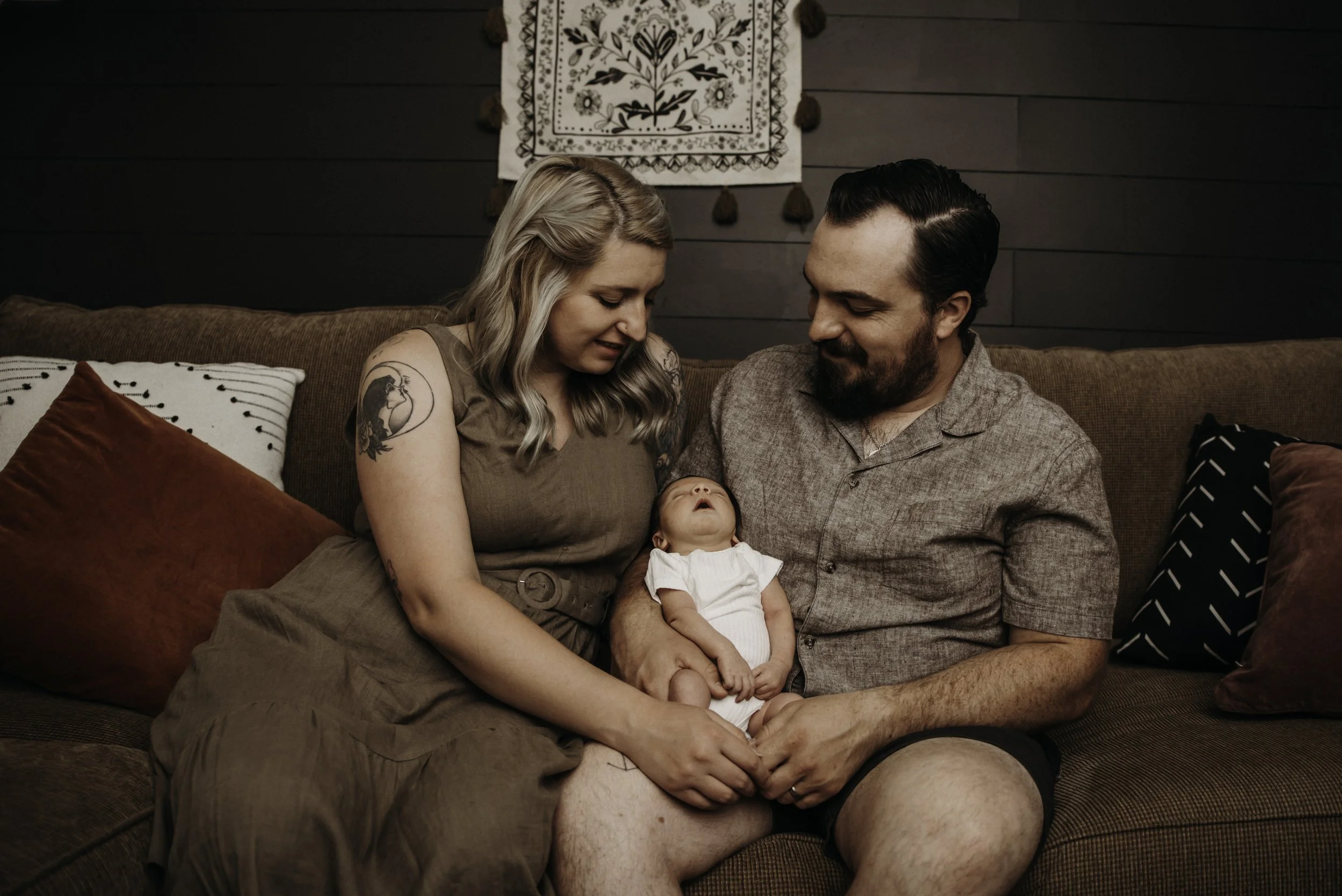 A family of three sitting on a brown couch, with a woman, a man, and a baby in a cozy living room. The woman has blonde hair and tattoos on her arm, the man has dark hair and a beard, and the baby is wearing a white outfit. They are looking at each other and the baby, creating a warm and affectionate scene.