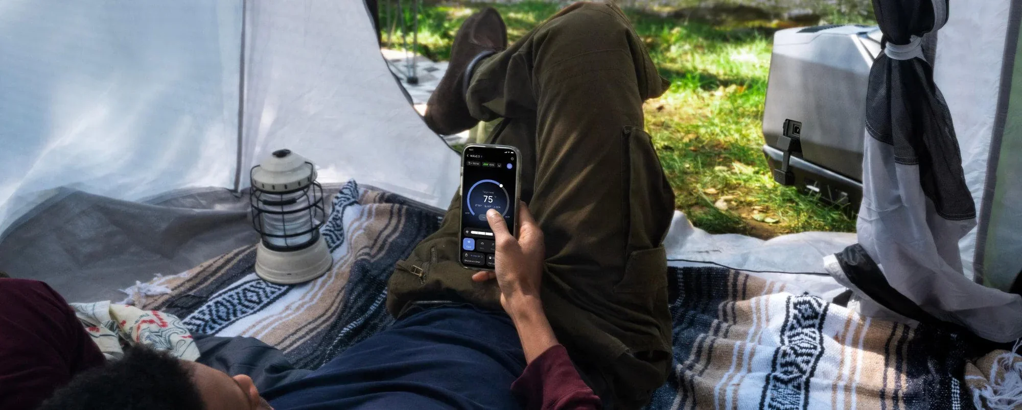 Person lying inside a tent using a phone to monitor temperature, surrounded by camping gear including a lantern and blankets, with grass and trees visible outside.