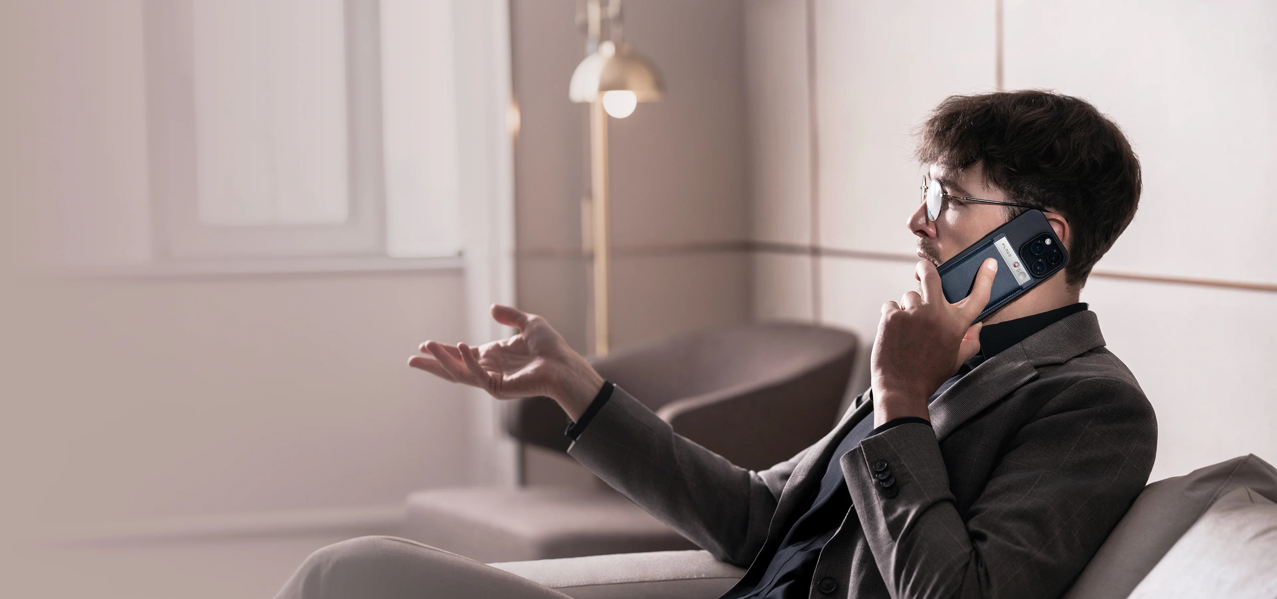 A young man with glasses and wearing a suit jacket is sitting on a sofa in a modern room, talking on a cell phone while gesturing with his left hand.
